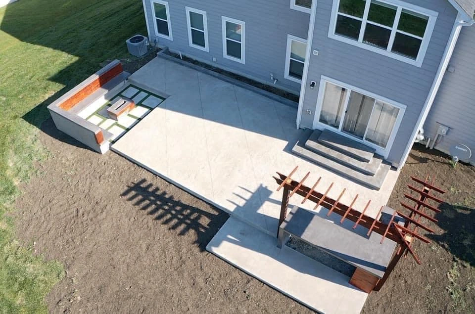 Aerial view of a modern backyard with a large concrete patio, steps to a gray house, and a wooden pergola over a sunken area.