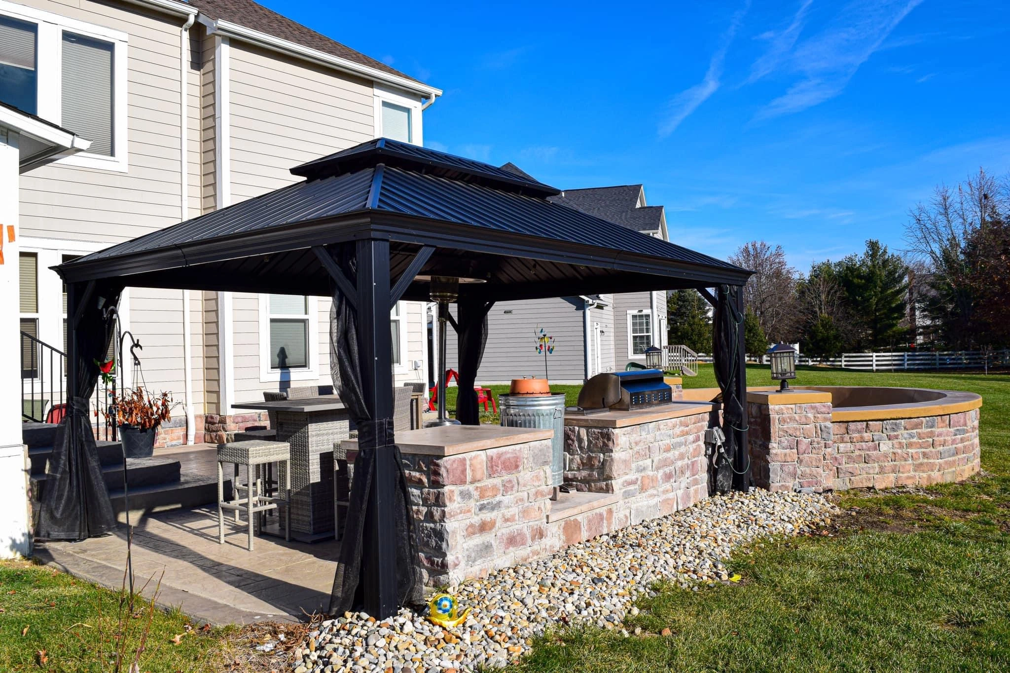 Backyard scene with a beige house, a black metal gazebo, outdoor seating, and a grill on a stone patio under a blue sky.