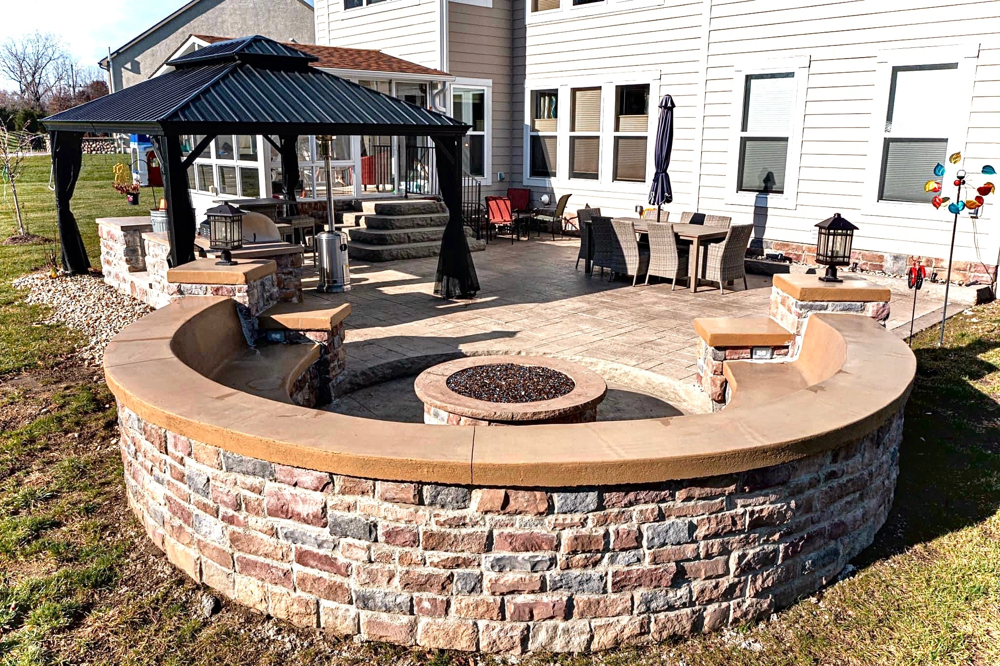 Circular stone fire pit wall surrounds a sunny backyard patio with wicker dining set and a metal-roofed gazebo beside a beige house.