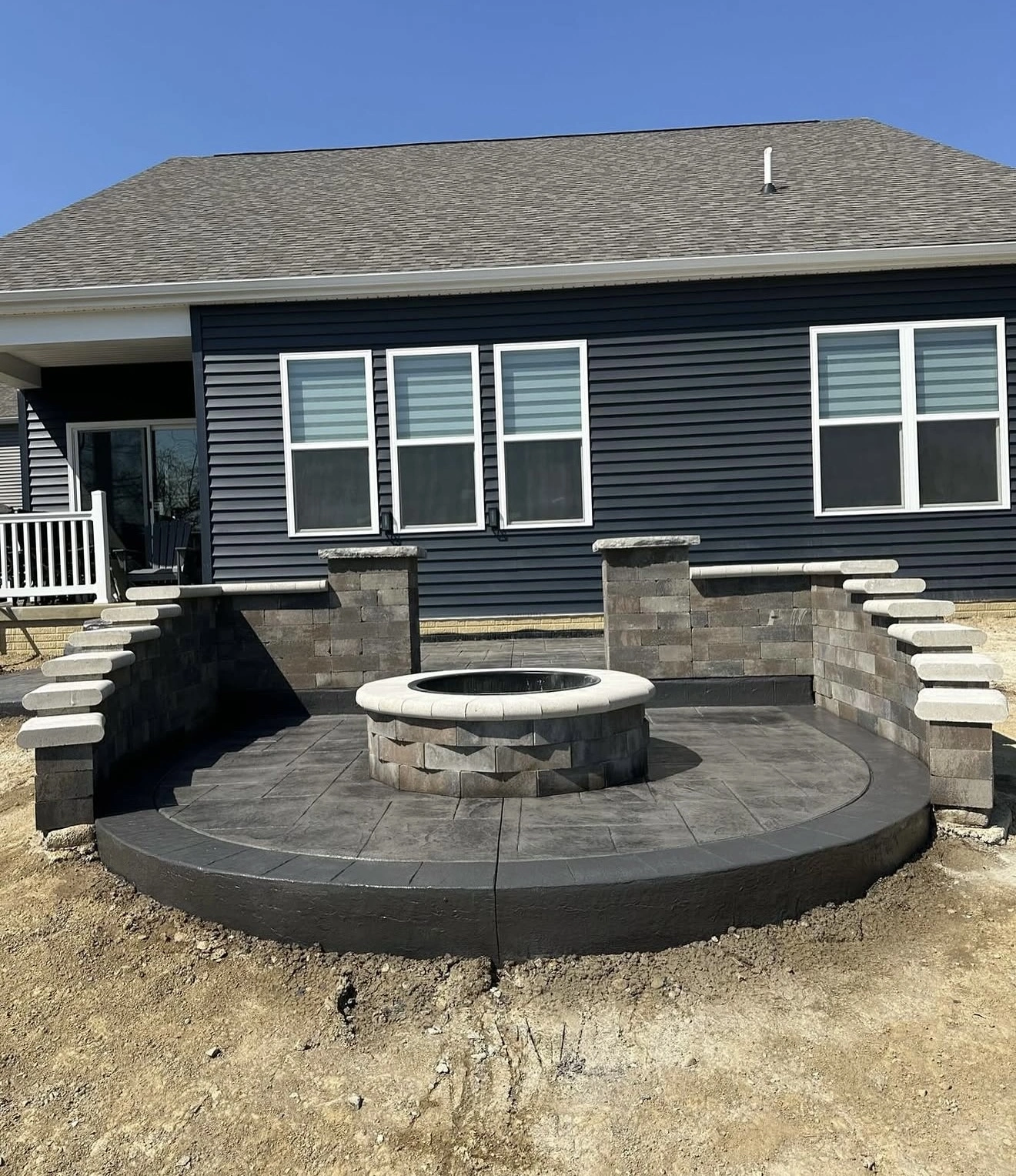 Circular stone patio with a central round fire pit, surrounded by short stone walls and stepped seating, in front of a dark blue house with white-framed windows under a clear blue sky.