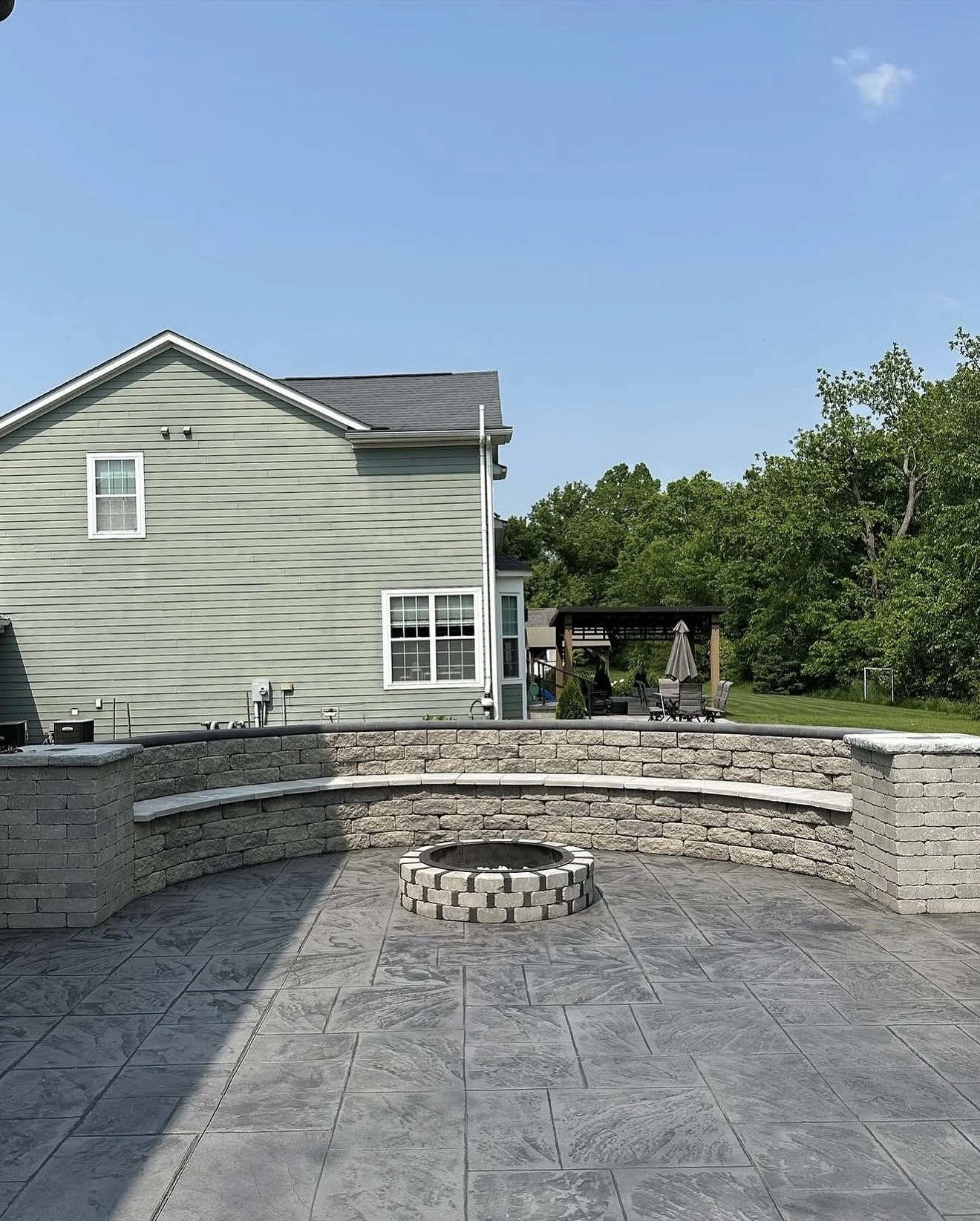 Backyard patio with a circular stone fire pit, curved seating wall, and a green-siding house in the background.