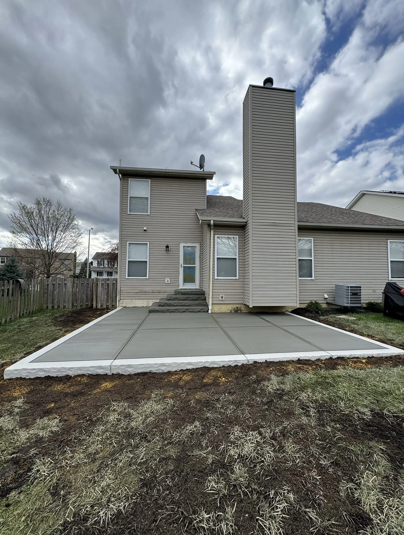 Beige vinyl-sided house with a tall central chimney, entry steps, and a large concrete patio on a cloudy day.