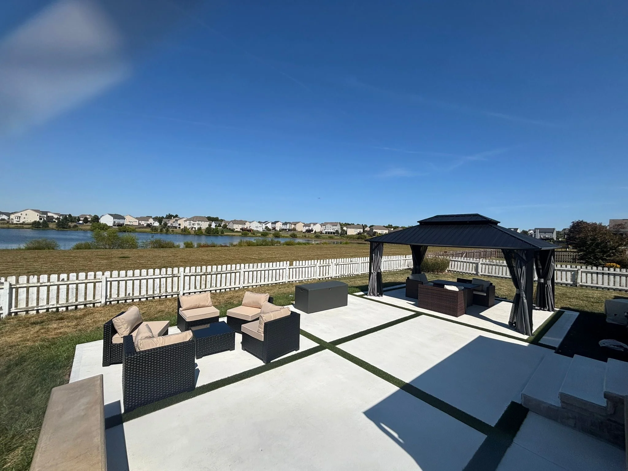 Outdoor backyard patio with wicker seating, a gazebo, and a white fence overlooking a lake and distant houses on a sunny day.