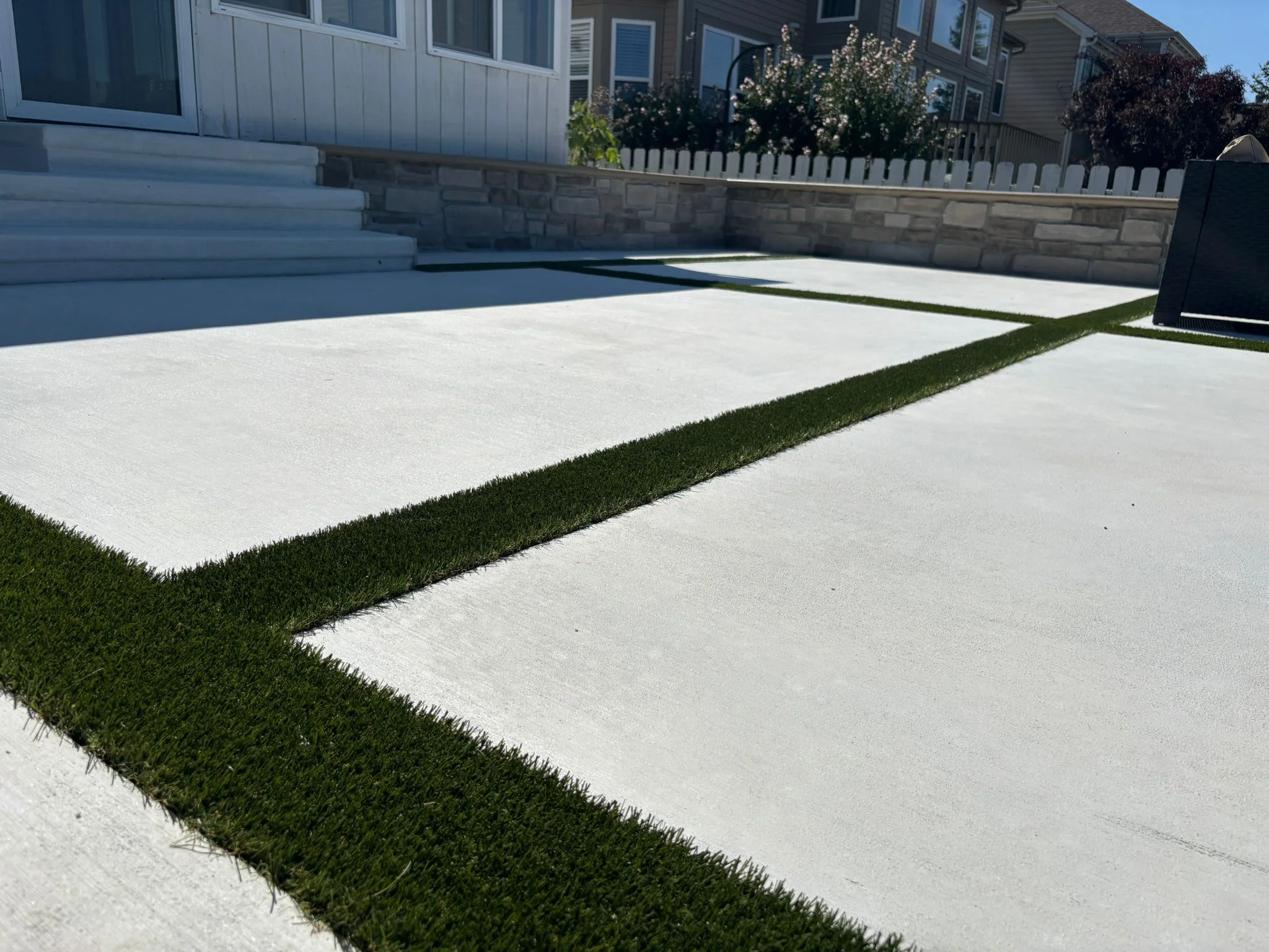Wide concrete driveway with narrow green artificial-turf strips between slabs, in front of a house with stone retaining wall and white fencing.