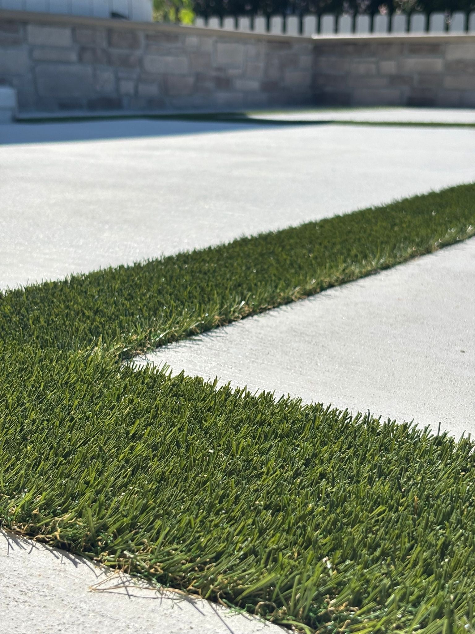 Low-angle view of artificial grass strips embedded in a concrete walkway along a low stone wall backdrop