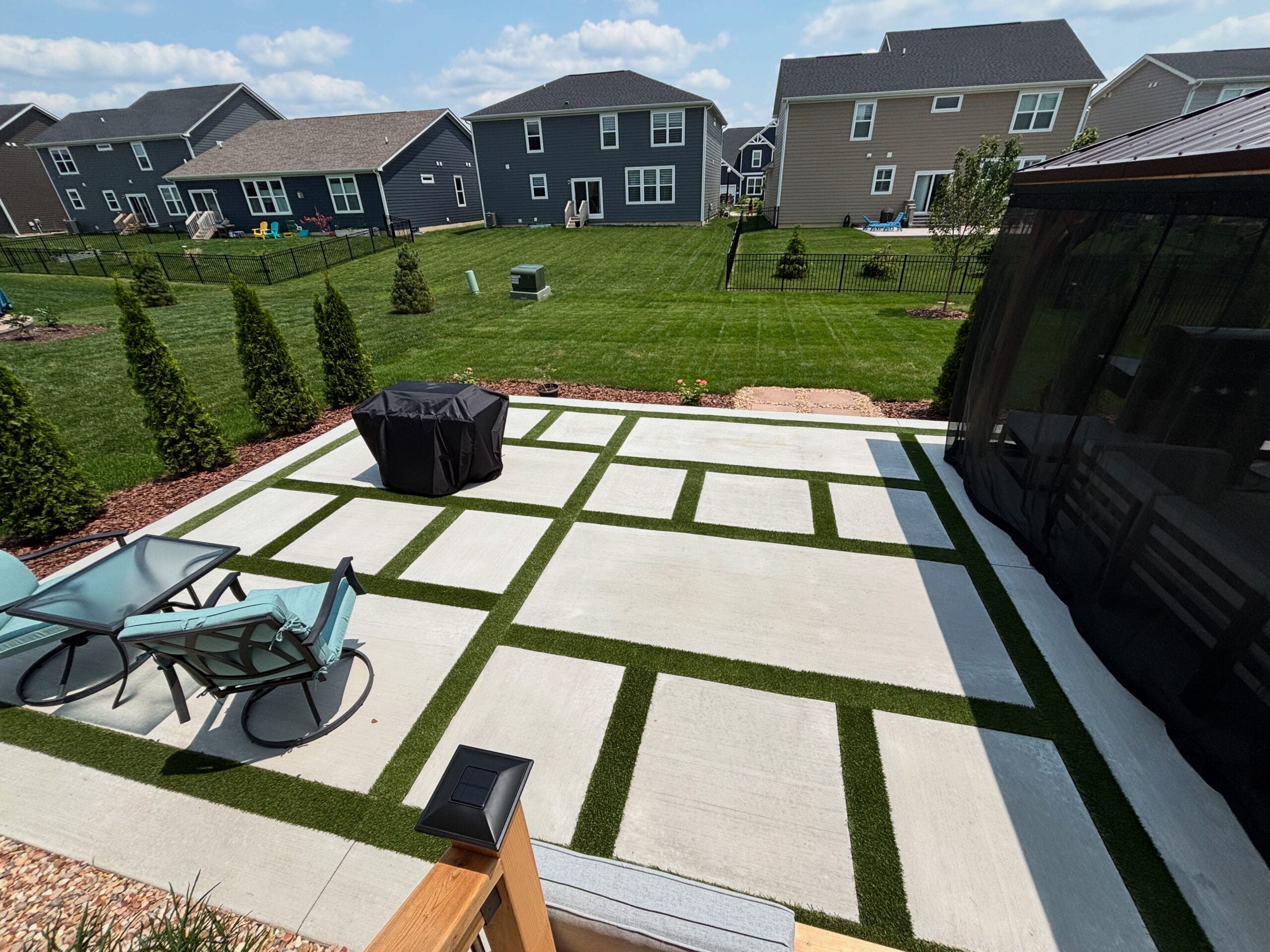 Backyard with geometric concrete patio and green turf strips, row of townhouses beyond, covered grill in foreground.