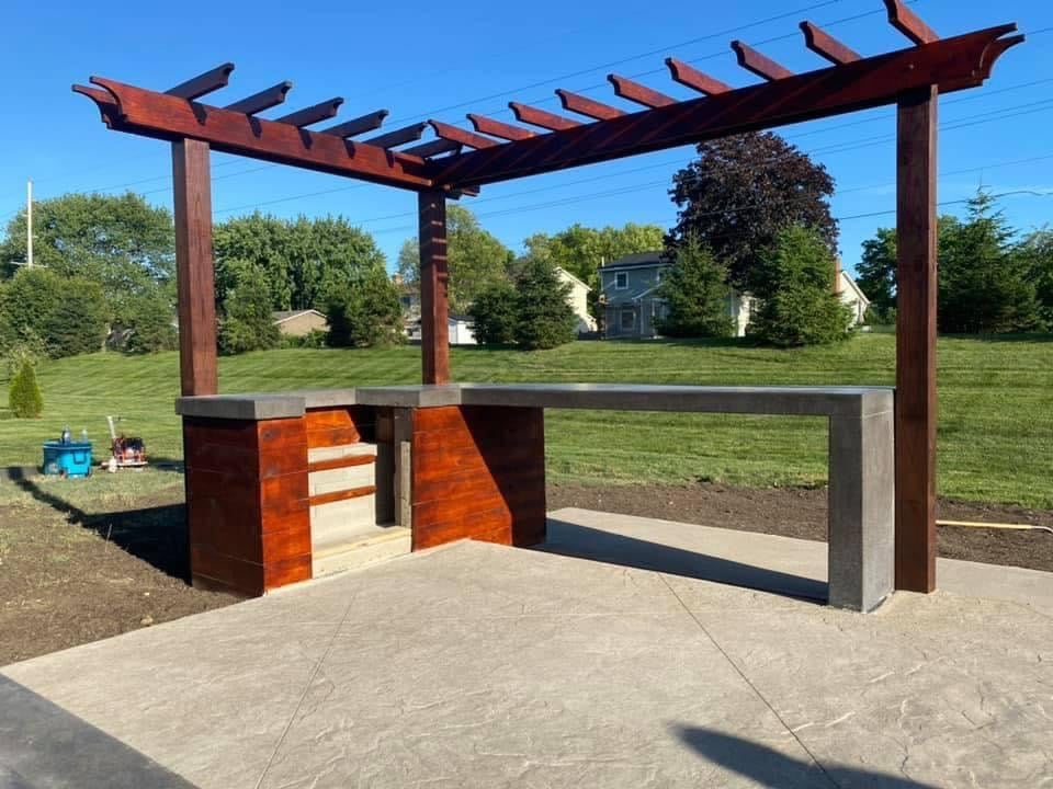 Outdoor outdoor kitchen/barbecue area with wooden pergola and concrete countertops, set on a paved patio.