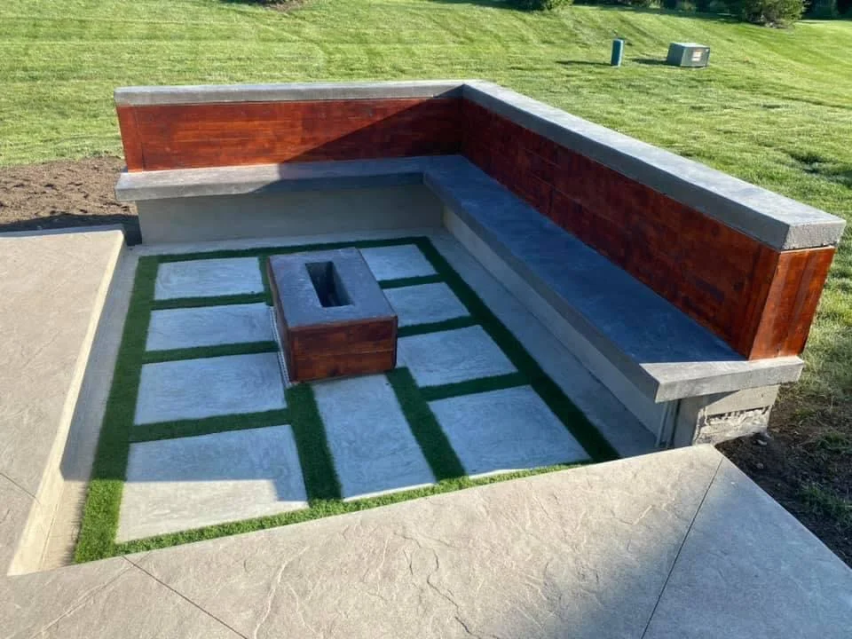 Built-in concrete patio with an L-shaped bench and a central square fire pit, framed by wood-panel walls.
