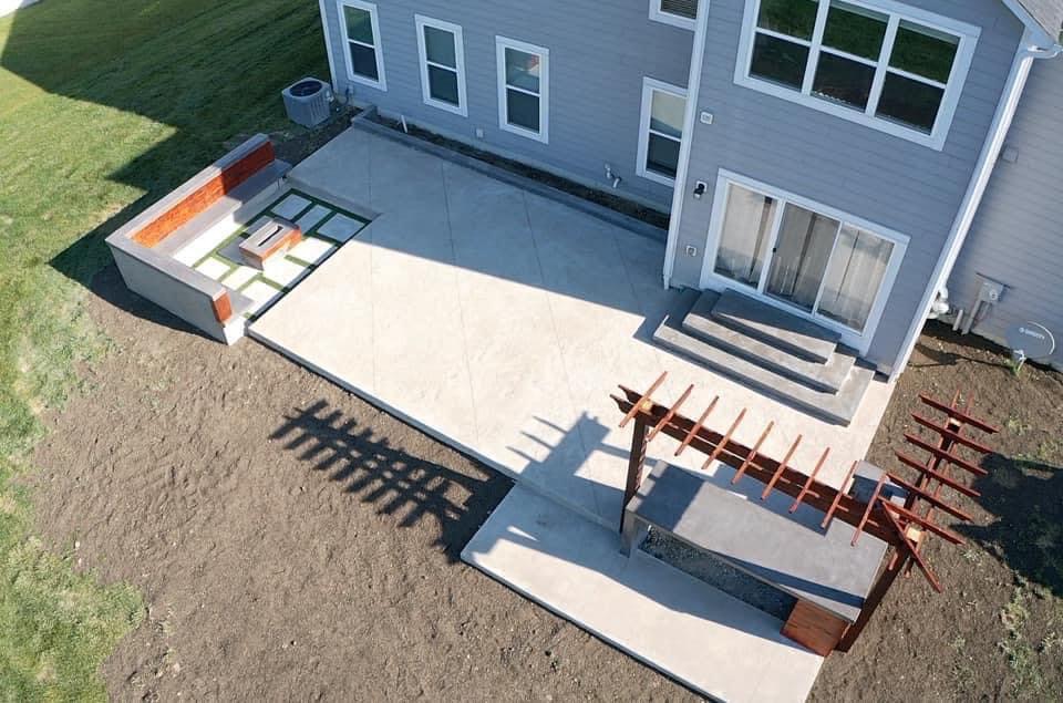 Aerial view of a grey two-story house with a large concrete patio, built-in brick seating, and a wooden pergola frame over a lower terrace area.