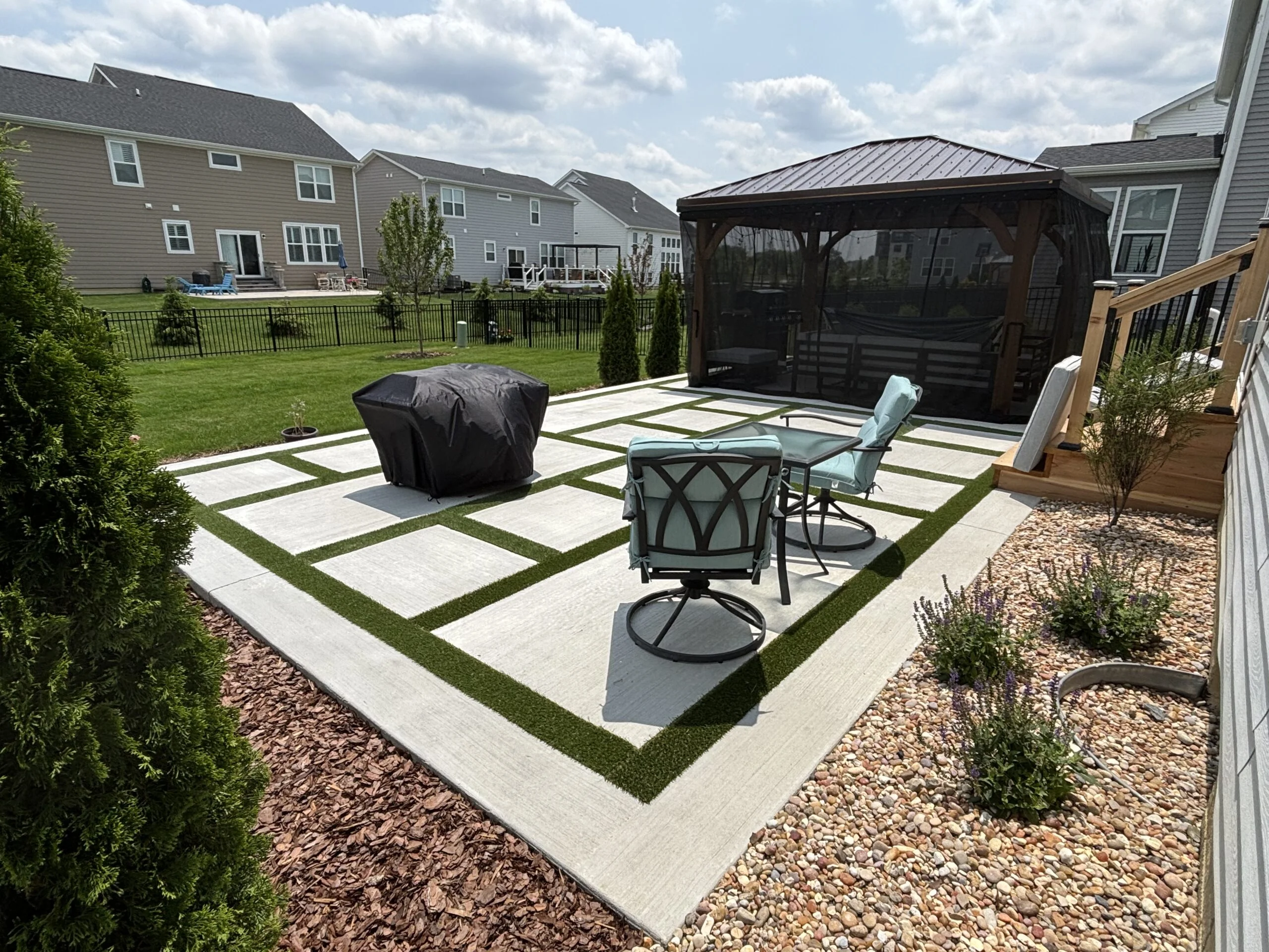 Backyard patio with a checkerboard of concrete squares and grass strips, outdoor furniture, and a covered grill.