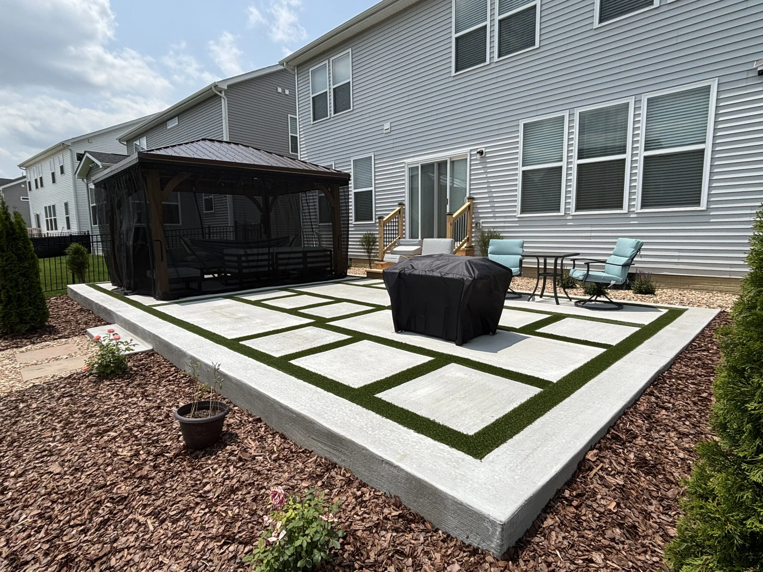 Backyard patio with concrete steps and artificial turf strips, a covered pergola, and outdoor seating beside a gray house.
