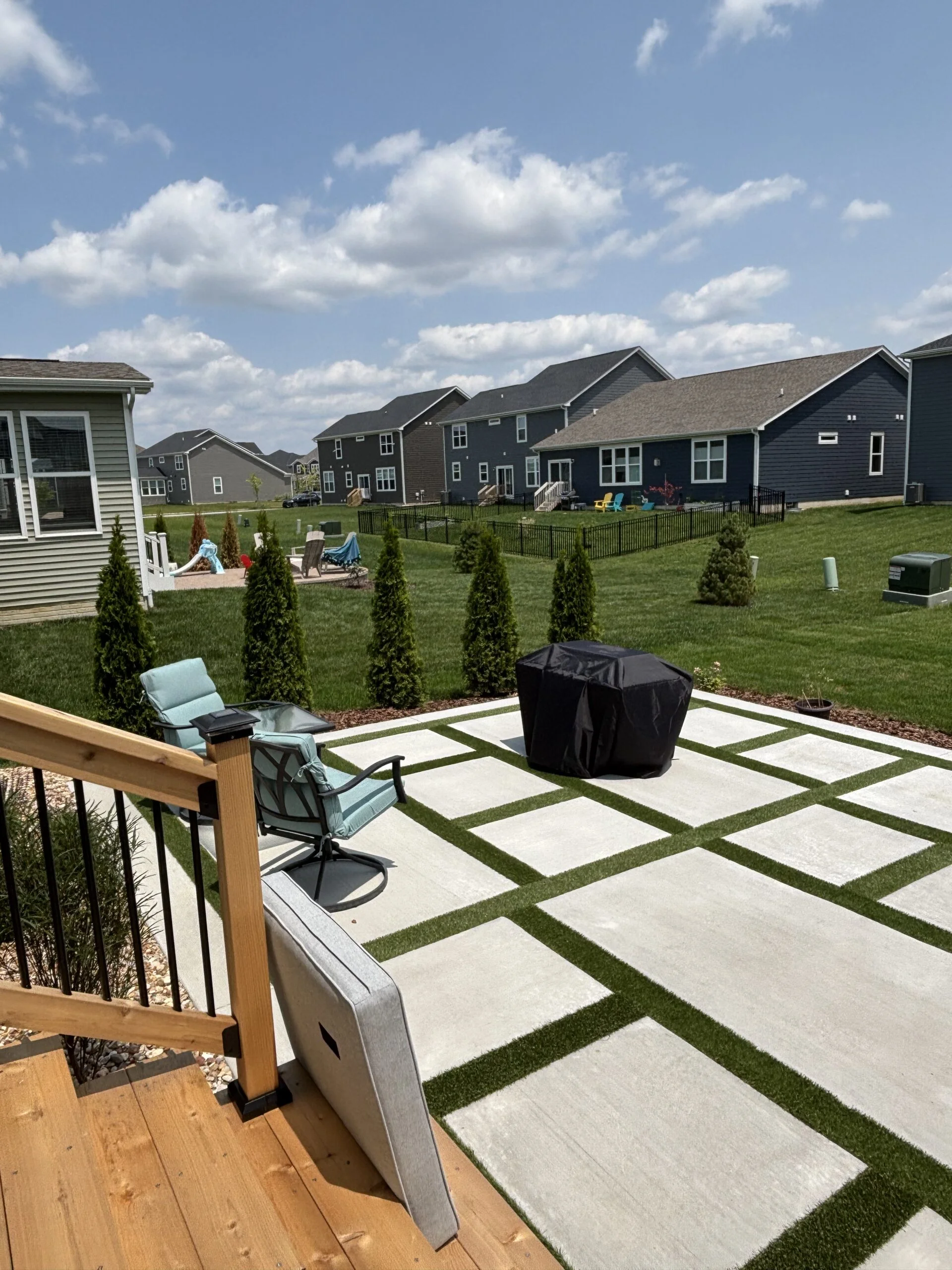 Backyard patio with large square concrete slabs separated by grass, a covered grill in the foreground and outdoor seating.