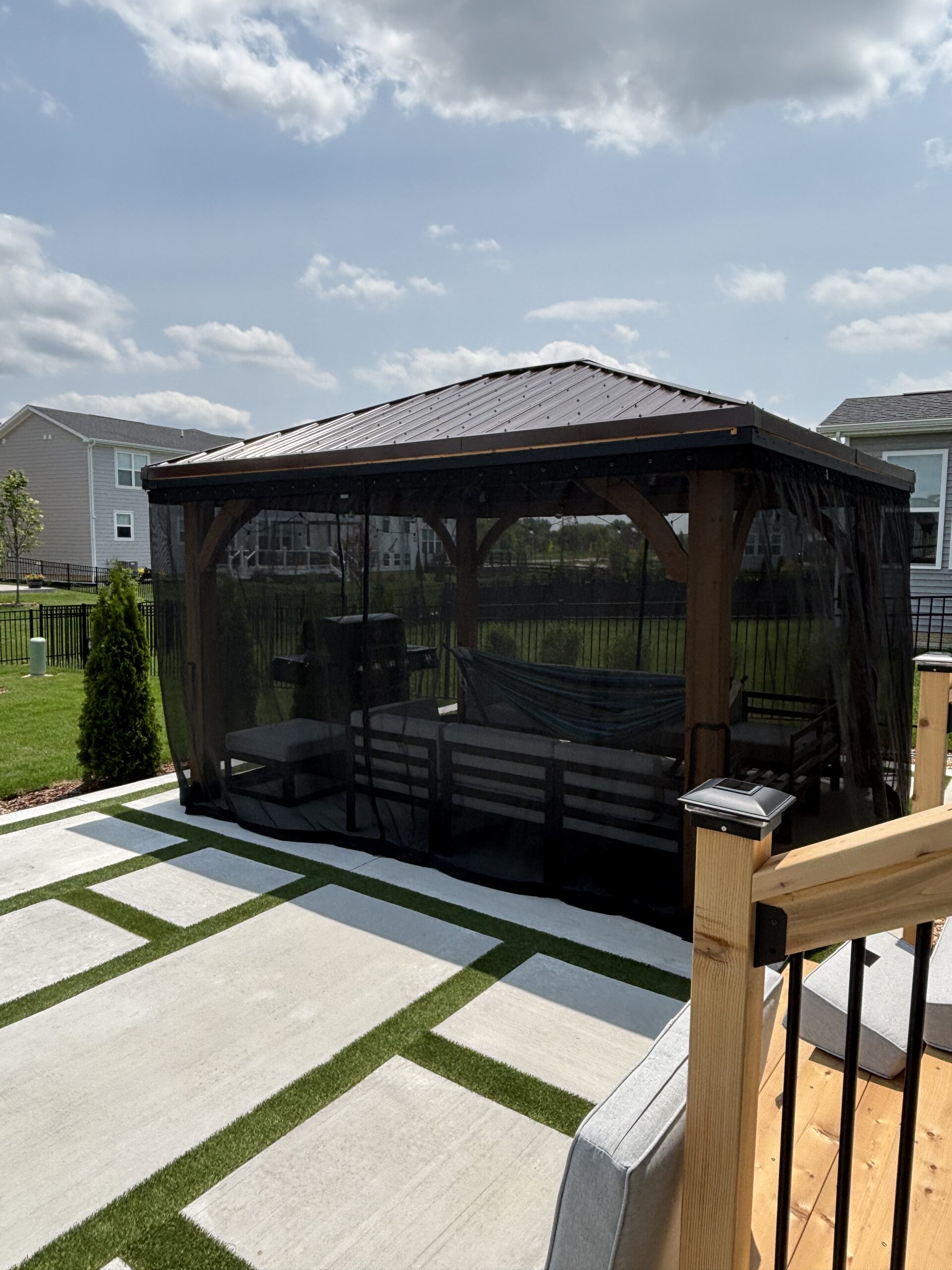 Backyard screened gazebo with seating under a metal roof, near suburban houses.