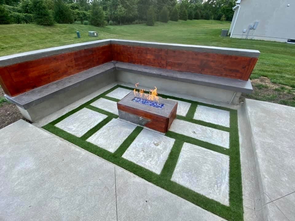 Modern outdoor fire area: rectangular fire pit on a raised concrete deck, flanked by wooden-paneled seating in a U‑shape with green turf squares nearby.