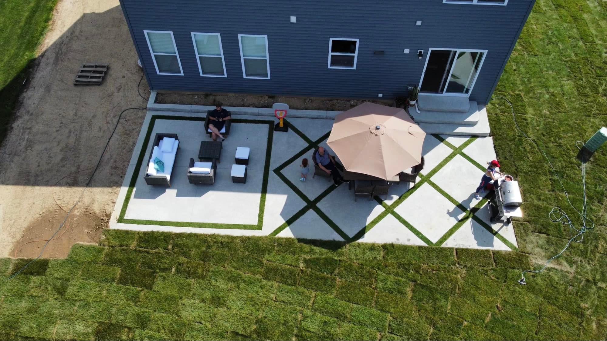Aerial view of a backyard patio beside a blue house with outdoor seating, a beige umbrella, and a grid-patterned concrete design outside. People are seated and standing near a grill area.