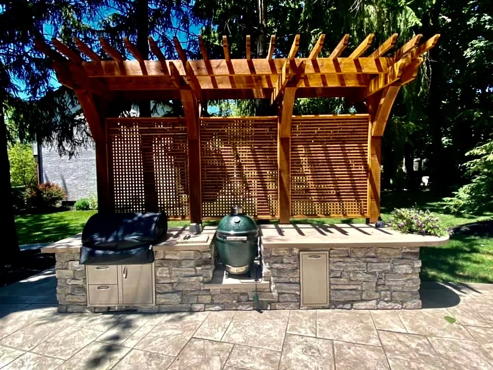 Outdoor stone kitchen counter with a built-in grill, a green ceramic smoker, and wooden pergola over lattice panels in a sunny backyard lighted scene.