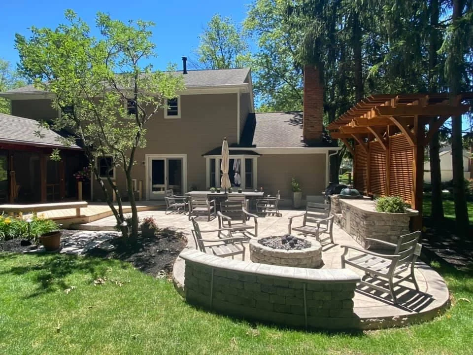 Backyard patio with a circular stone fire pit and surrounding metal chairs, in front of a grey house with a glass door and an umbrella-covered dining area.