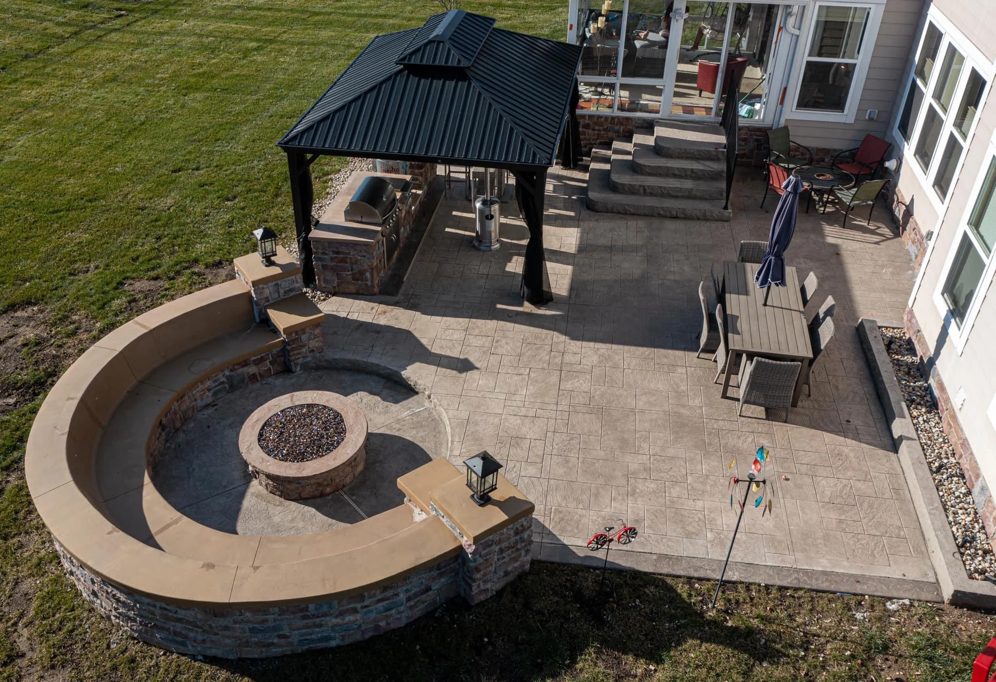 Aerial view of a circular stone fire pit with seating wall, a covered outdoor kitchen area, and a paved patio with dining table and chairs adjacent to a house.