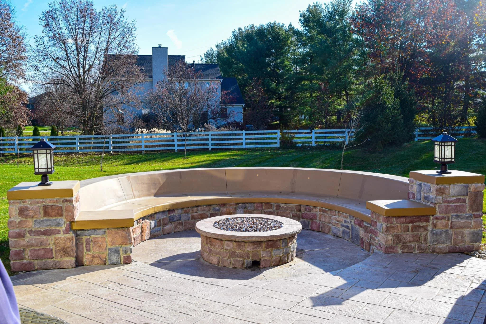 Circular stone patio with built-in seating surrounding a gravel-filled fire pit, two post lanterns, white fence, and a lawn with trees in the background on a sunny day.