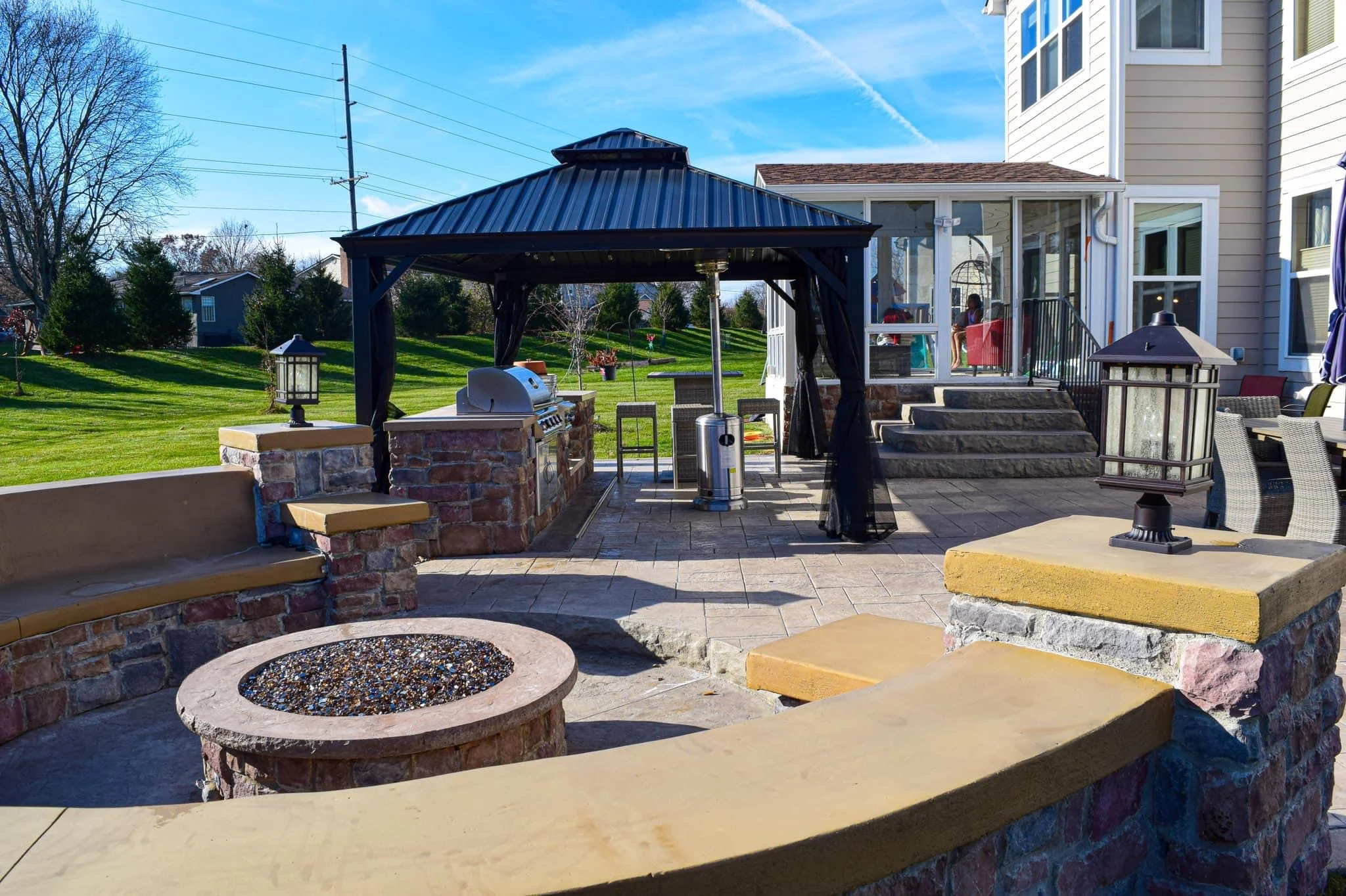 Backyard patio with a blue metal-roofed gazebo, built-in grill, stone planters, and seating beside a beige house.