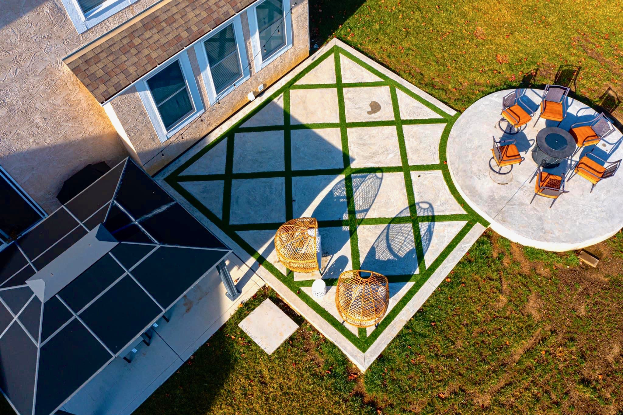 Aerial view of a modern backyard: beige house wall, geometric concrete patio with green turf grid, and a circular patio with orange-cushioned chairs around a table.