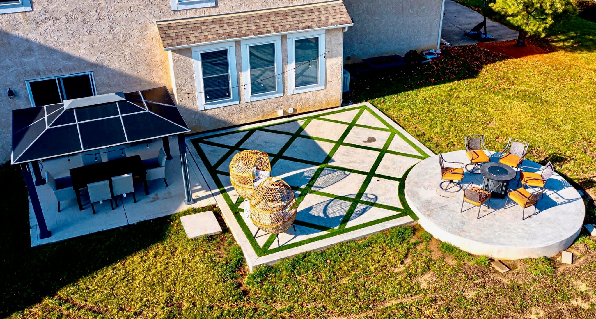 Aerial view of a backyard patio with a geometric turf-and-concrete pattern, a round seating deck, and a dining area under a covered patio.