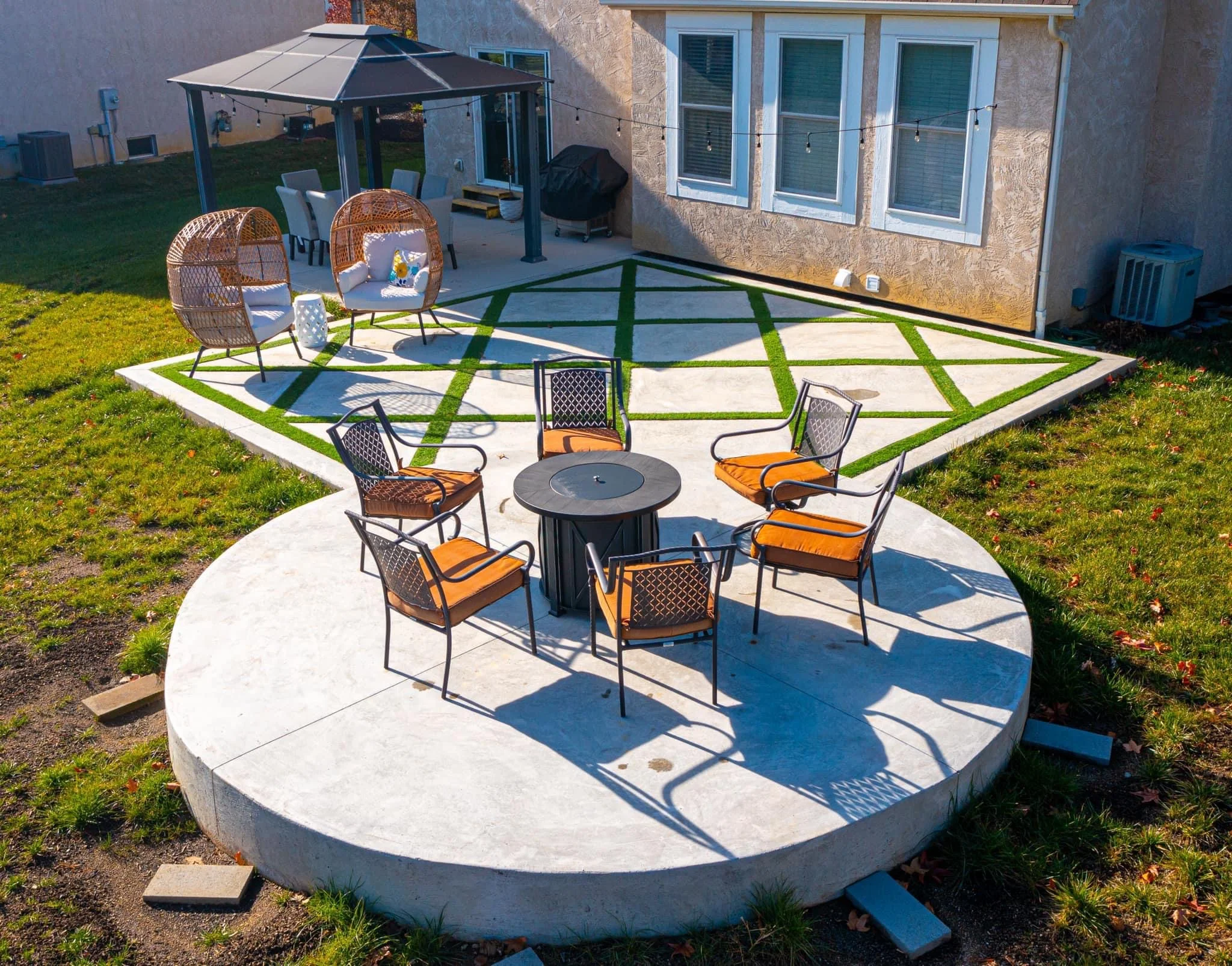 Circular concrete patio with a fire pit table and orange-cushioned chairs surrounding it, in a grassy backyard with a gazebo and string lights.