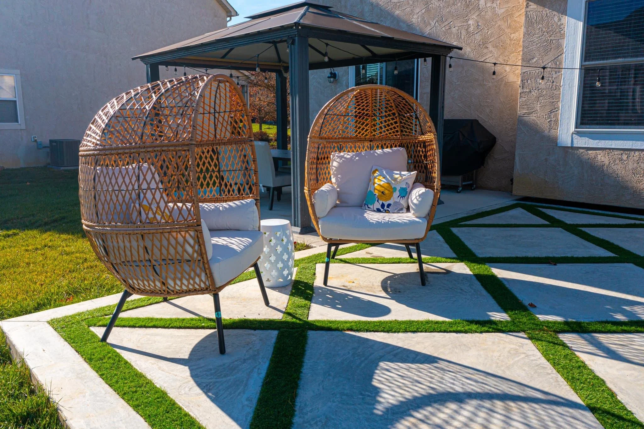 Outdoor patio scene with two woven dome chairs, cushions, and a small white table under a gazebo, overlooking a grassy backyard with string lights overhead.