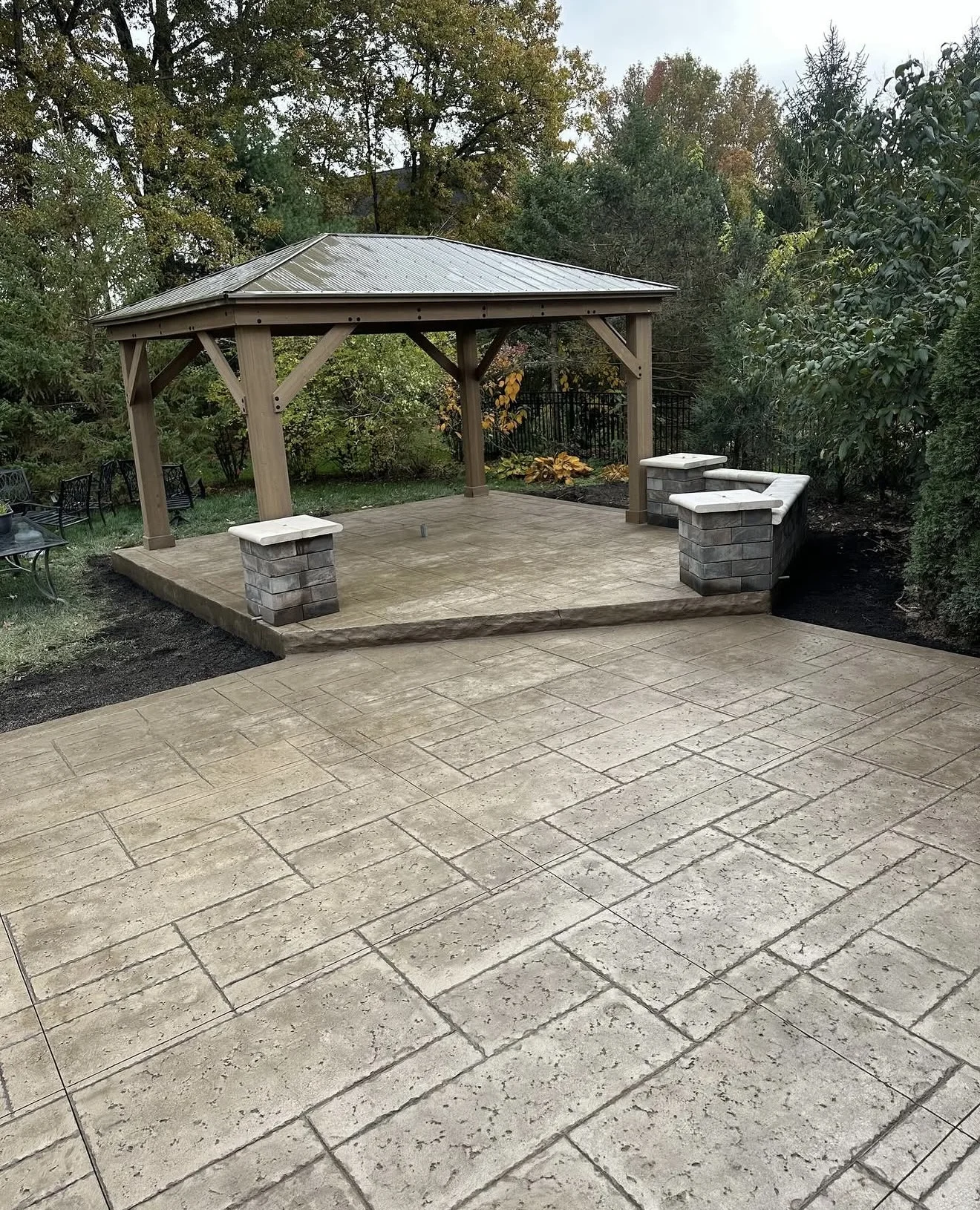 Wooden gazebo with a metal roof over a tiled backyard patio, surrounded by trees and shrubs.