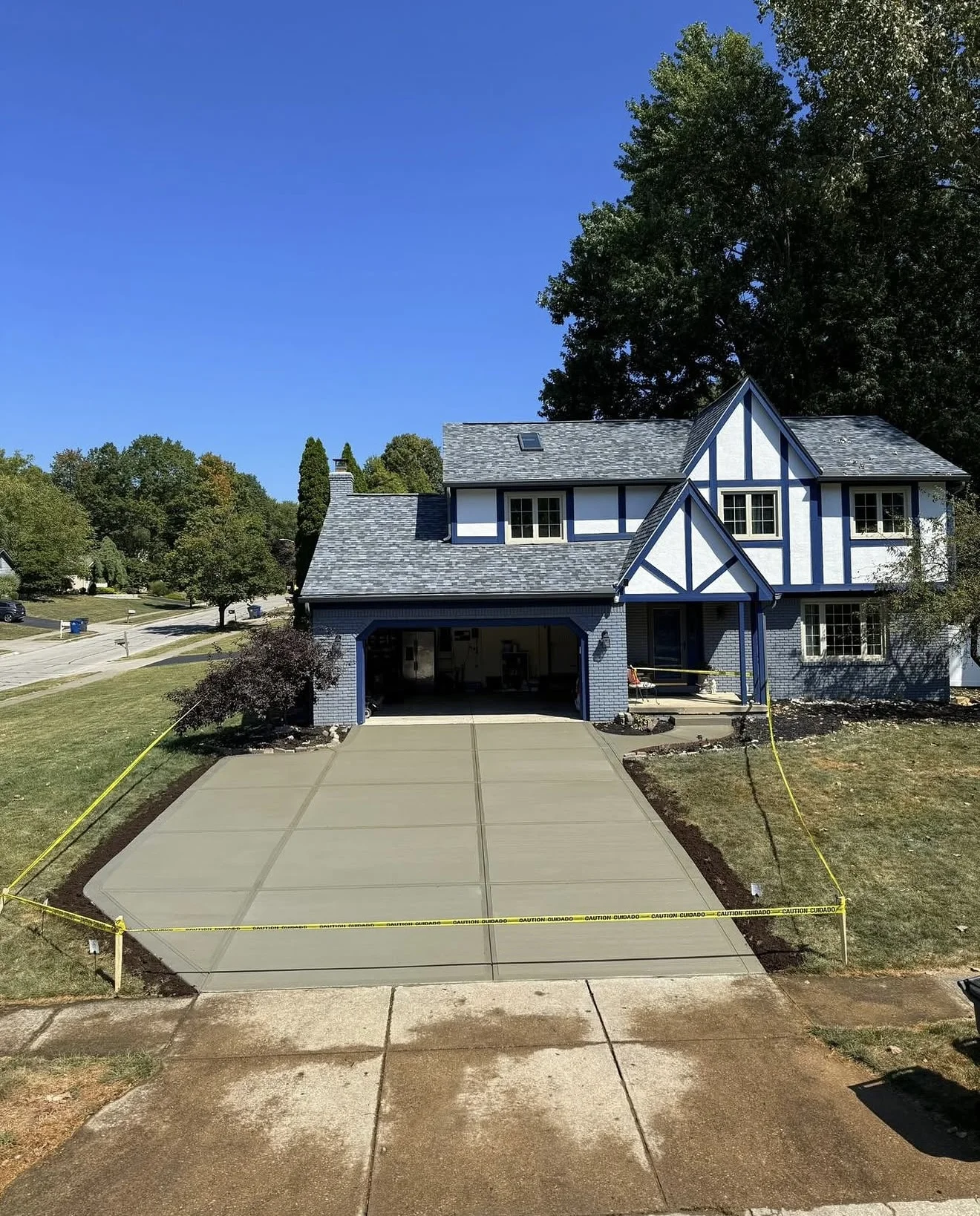 Two-story blue house with white trim and a newly poured wide concrete driveway, edged by yellow caution tape.