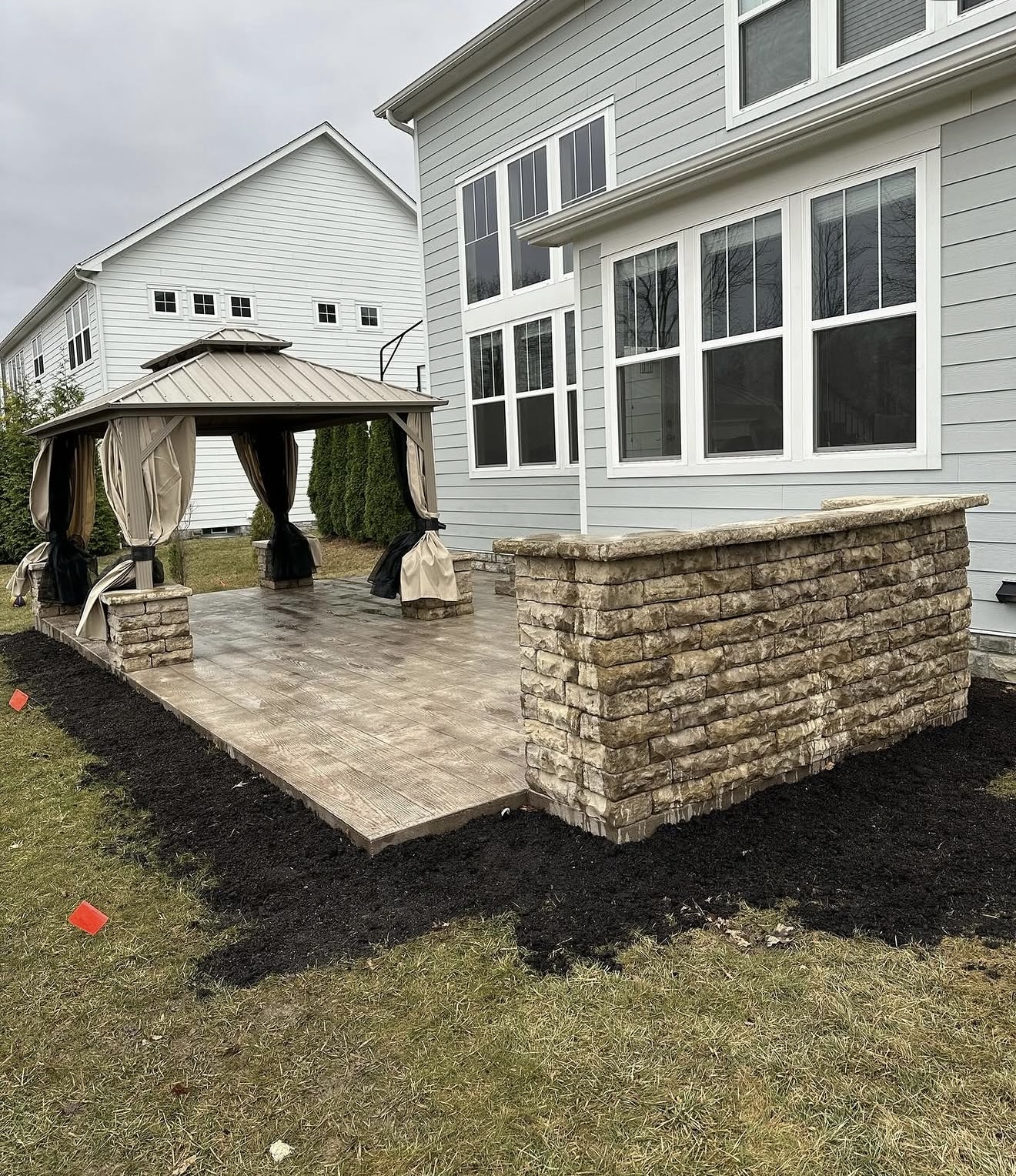 Outdoor patio with a beige gazebo and draped curtains on a stamped concrete slab beside a gray-siding house with large windows and a stone wall edge.