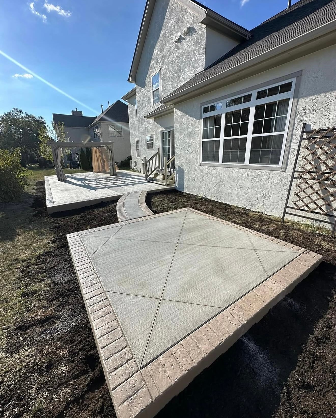 Backyard patio area with a large raised concrete deck, a pergola, and a white stucco house along with a curved stepping path.