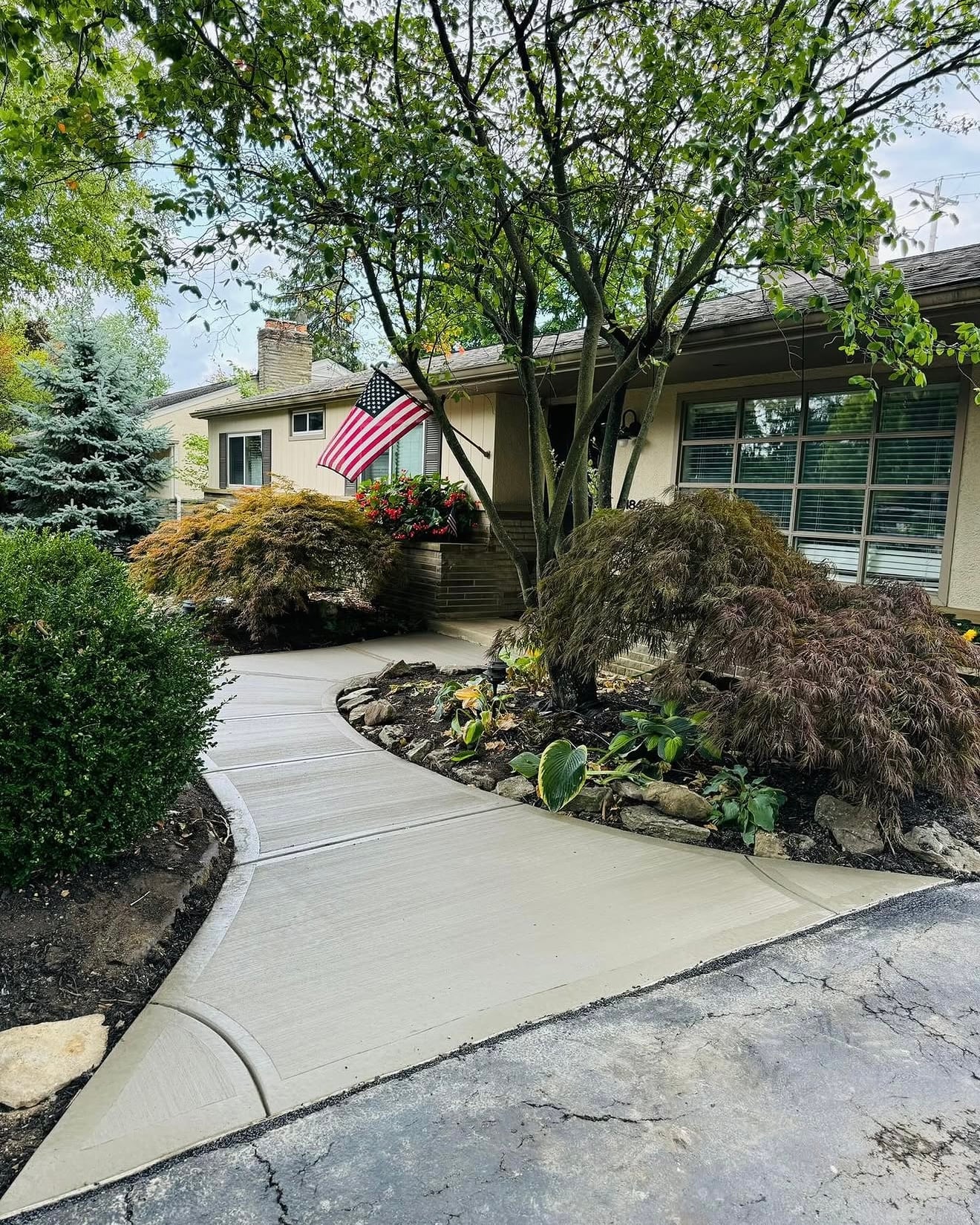 Front yard of a suburban house with a curved concrete walkway, trees, and an American flag by the steps.