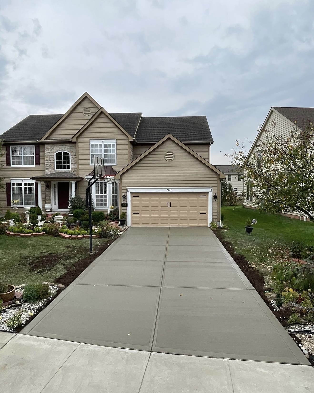 Two-story beige house with a double garage, basketball hoop, and landscaped front yard guarded by a small garden bed.