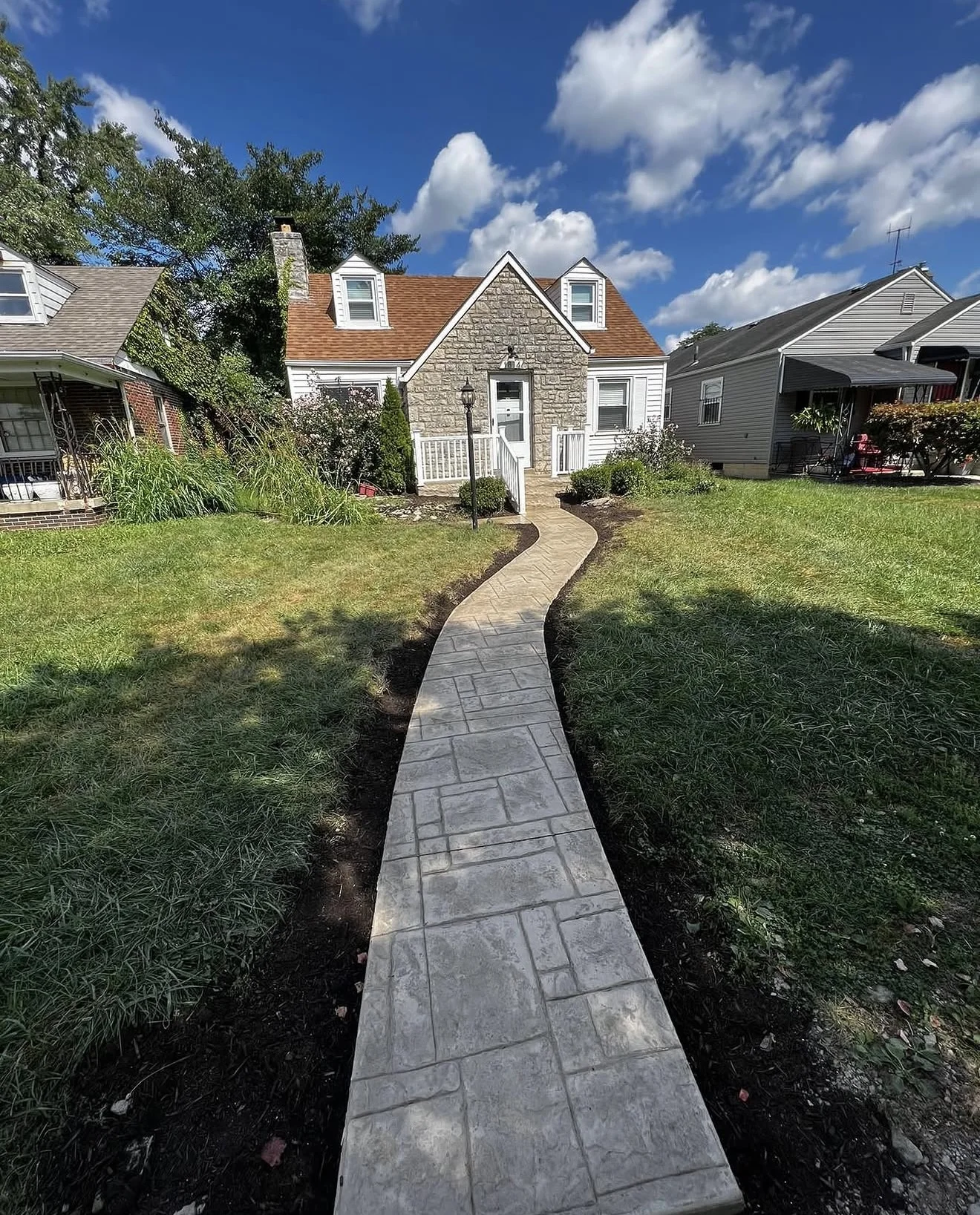 Curved stone walkway leads to a small stone-and-siding cottage with white trim under a bright blue sky.
