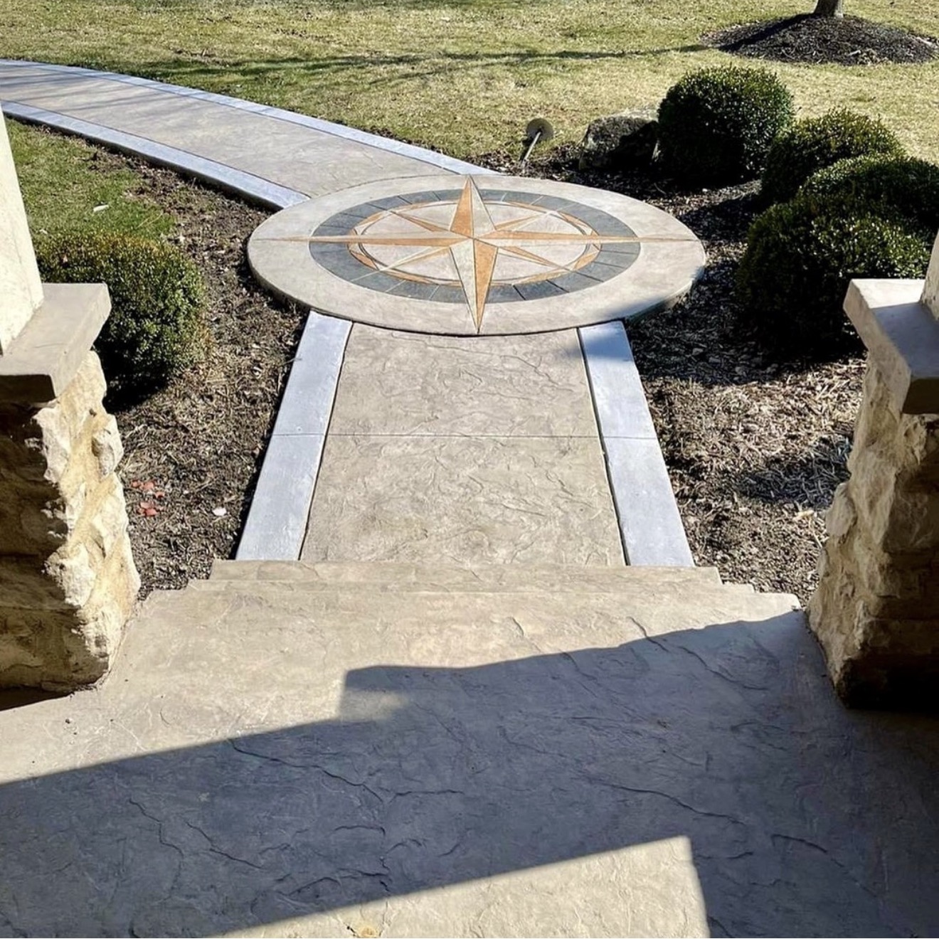 Circular compass rose stone feature at end of a stone walkway, with surrounding landscaped bushes and steps in the foreground.