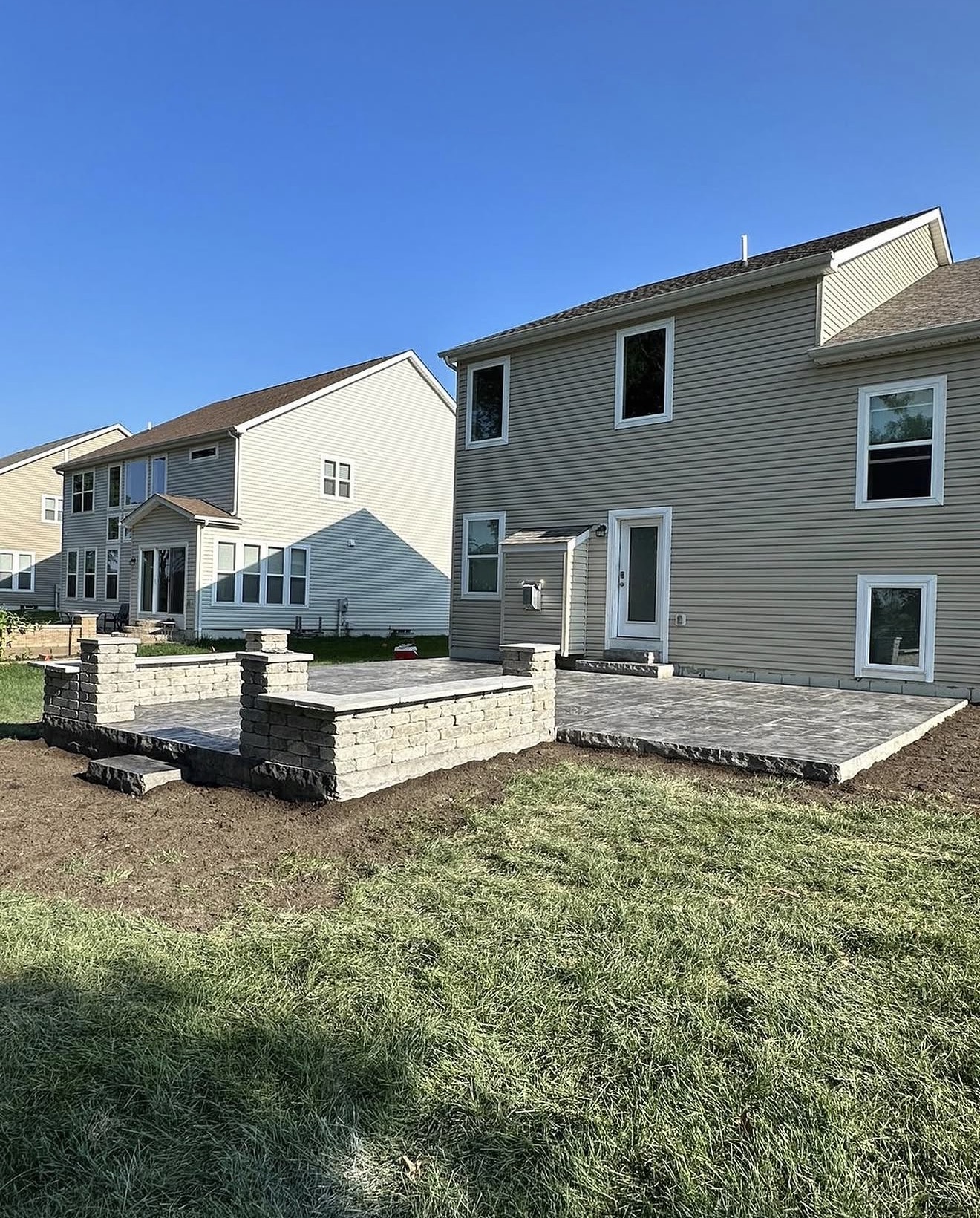 New two-story beige house with gray siding and a stone patio under a clear blue sky, in a housing development.