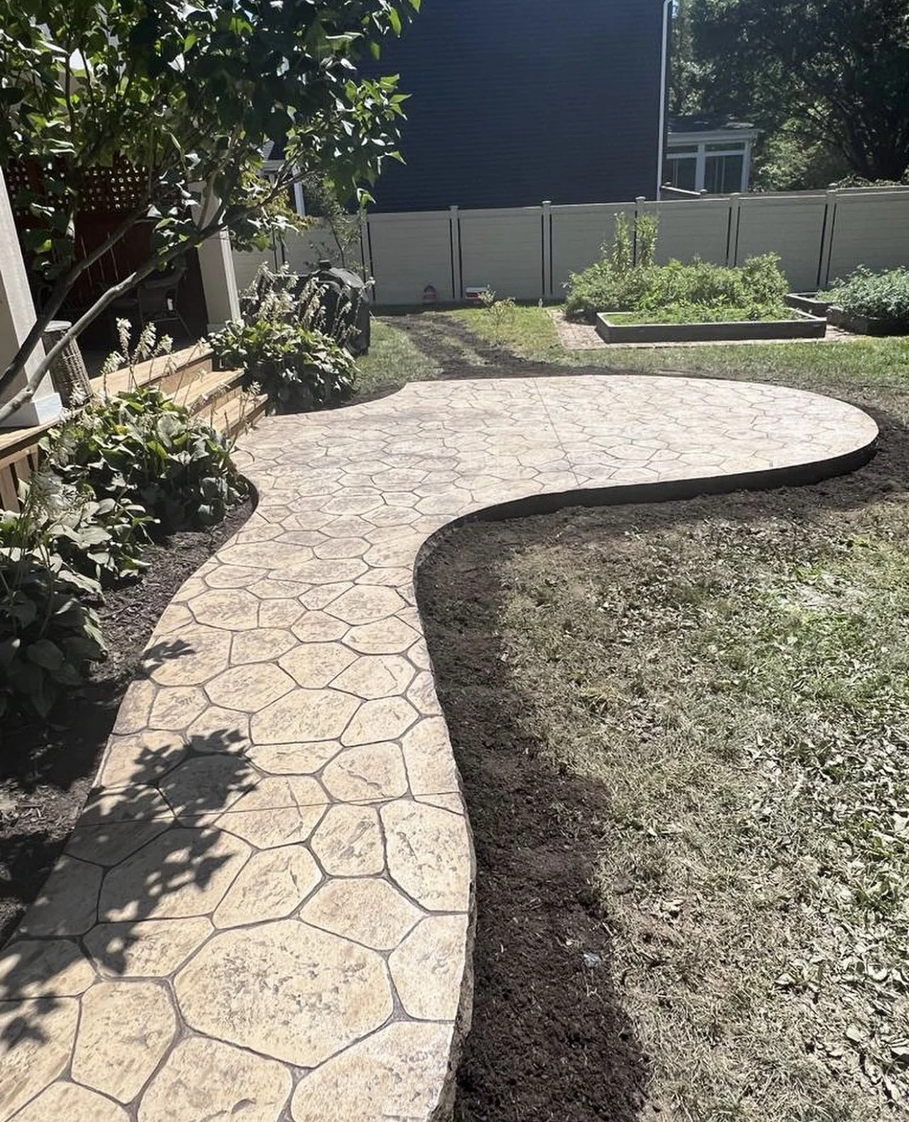Curved stamped-concrete backyard path with a stone pattern looping toward raised garden beds and a privacy fence nearby.