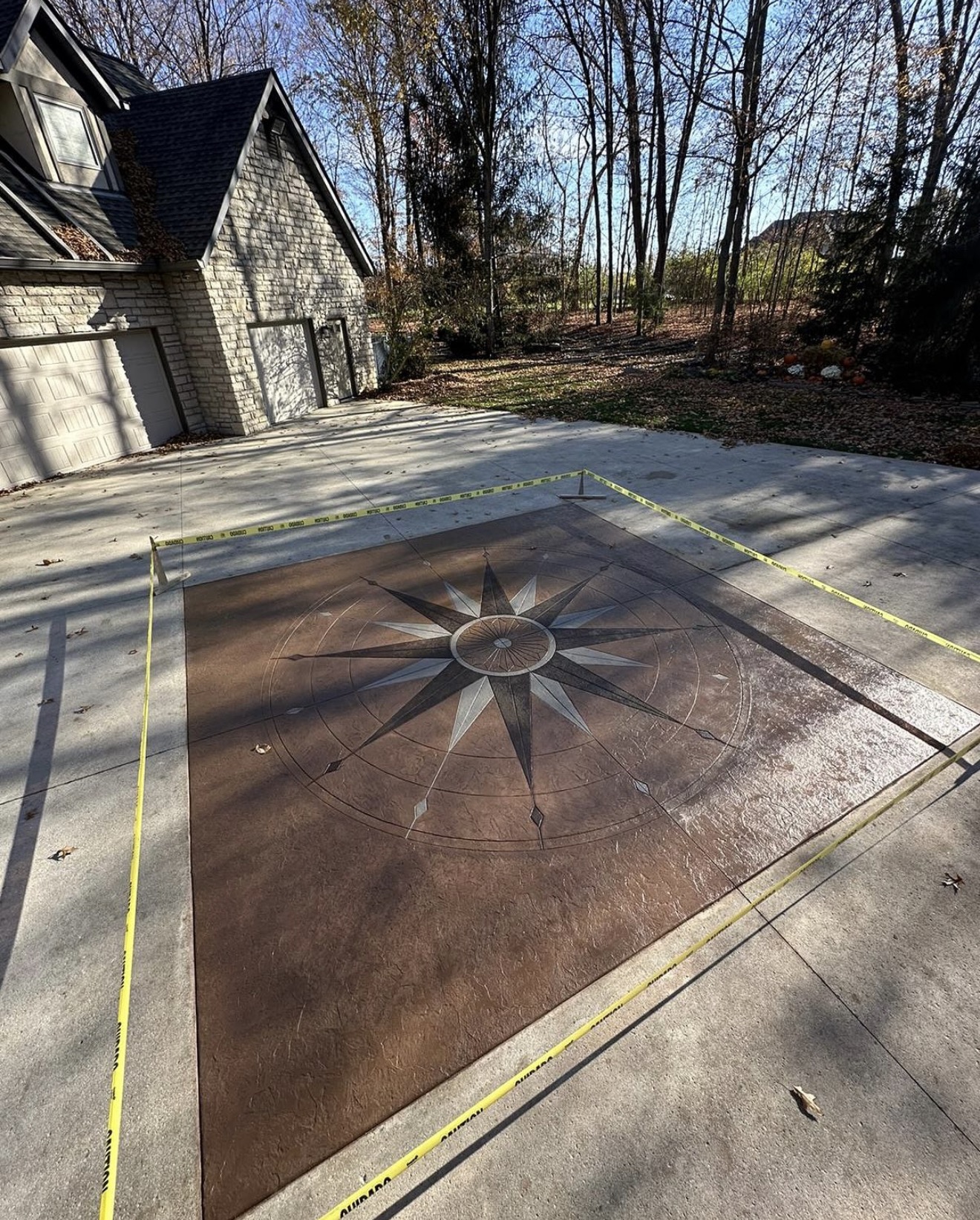 Decorative compass rose inlay on a concrete driveway, bordered by yellow caution tape, with a stone-clad house and leafless trees in the background.