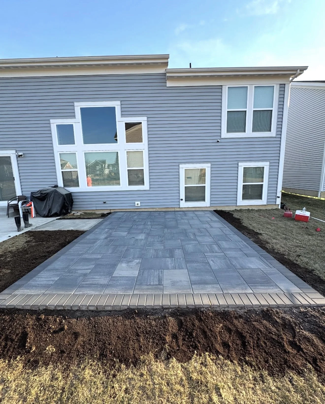 Backyard of a blue house with a large window wall and a newly laid gray tiled patio, dirt edging along the border.