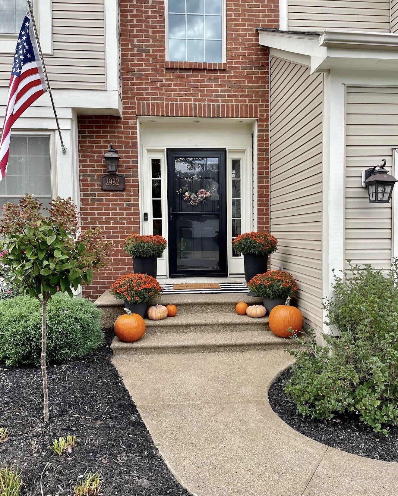 Front porch of a brick townhouse with a black front door, pumpkins and potted mums on the steps, and an American flag to the left.