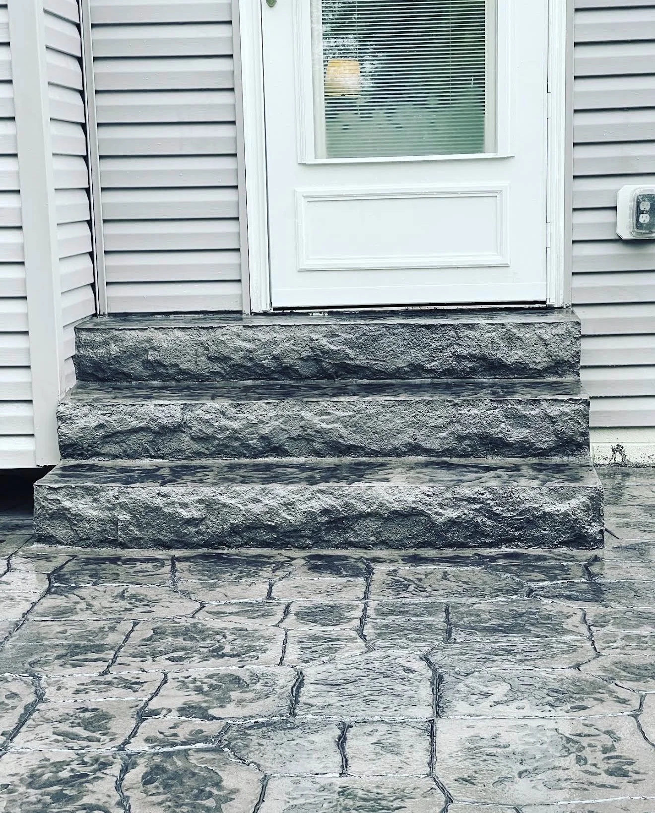Stone front steps leading to a white door with blinds, gray siding, and stone thresholds outside a home.