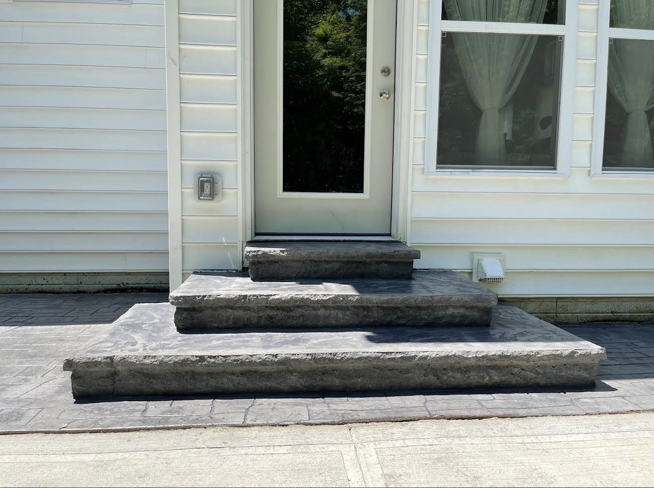Three-step concrete entry stoop leading to a light-colored door on a white-sided house, with two windows to the right covered by sheer curtains