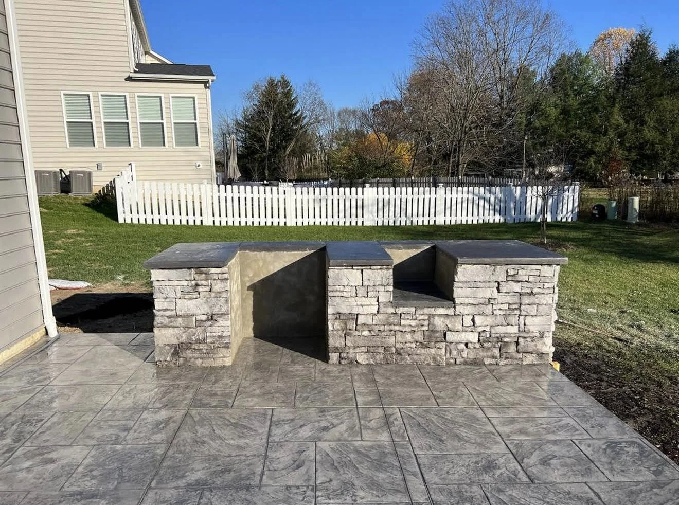 Outdoor stone counter/bar with three sections on a gray tiled patio, white picket fence and lawn behind.