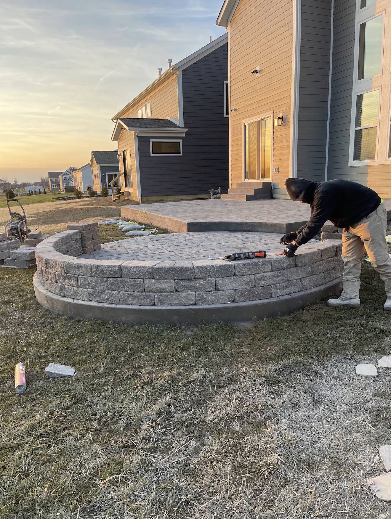 Worker in a hoodie building a circular stacked-stone wall around a raised patio next to a modern house at sunset.
