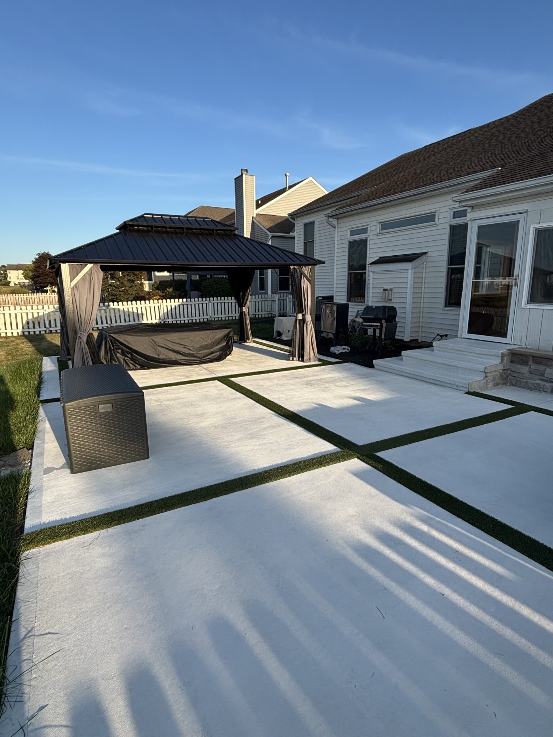 Backyard patio with a black gazebo, beige curtains, and white concrete slabs bordered by artificial grass.