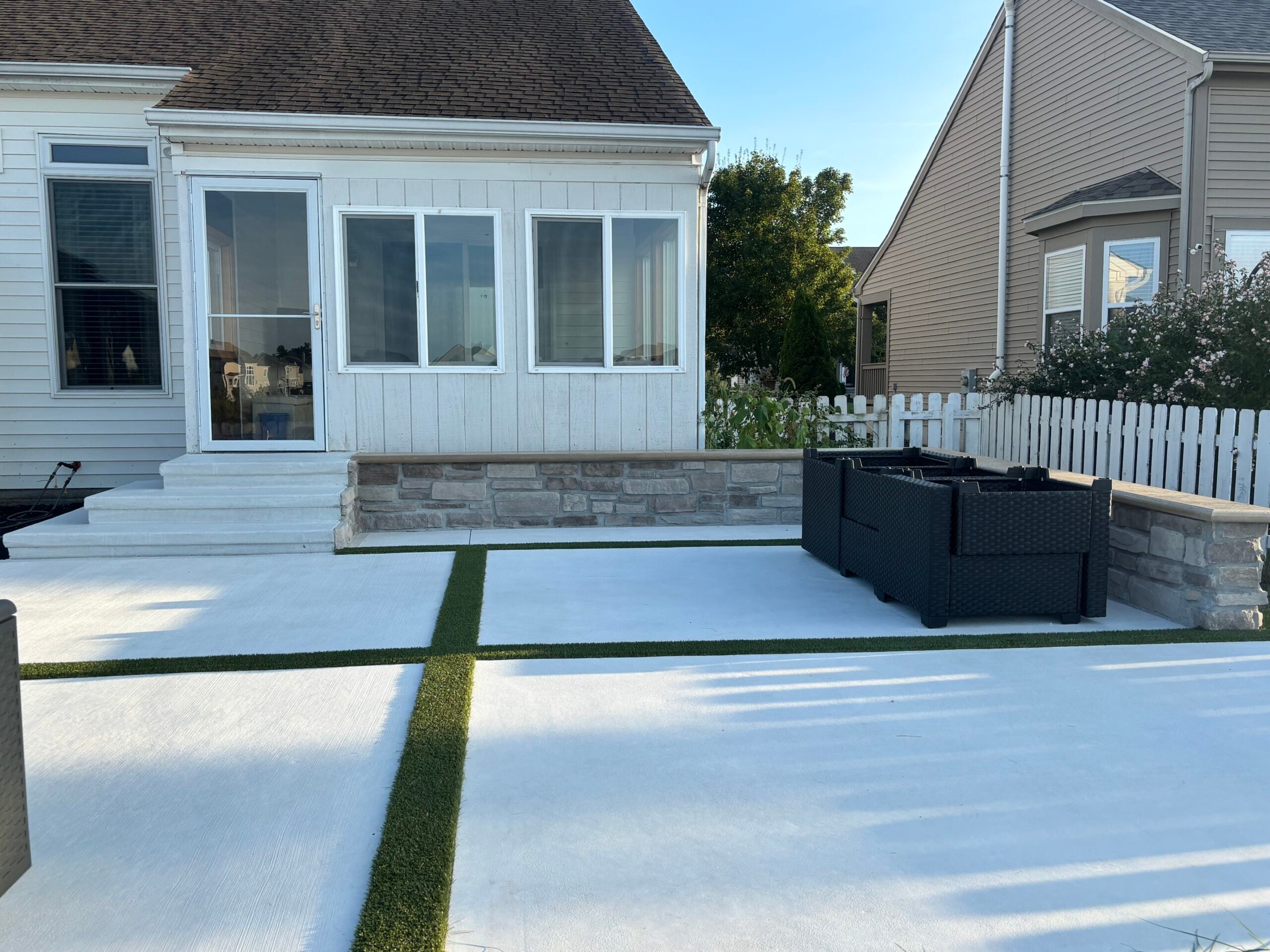 Backyard patio with white concrete slabs and narrow artificial grass strips, glass doors and windows on a white house.