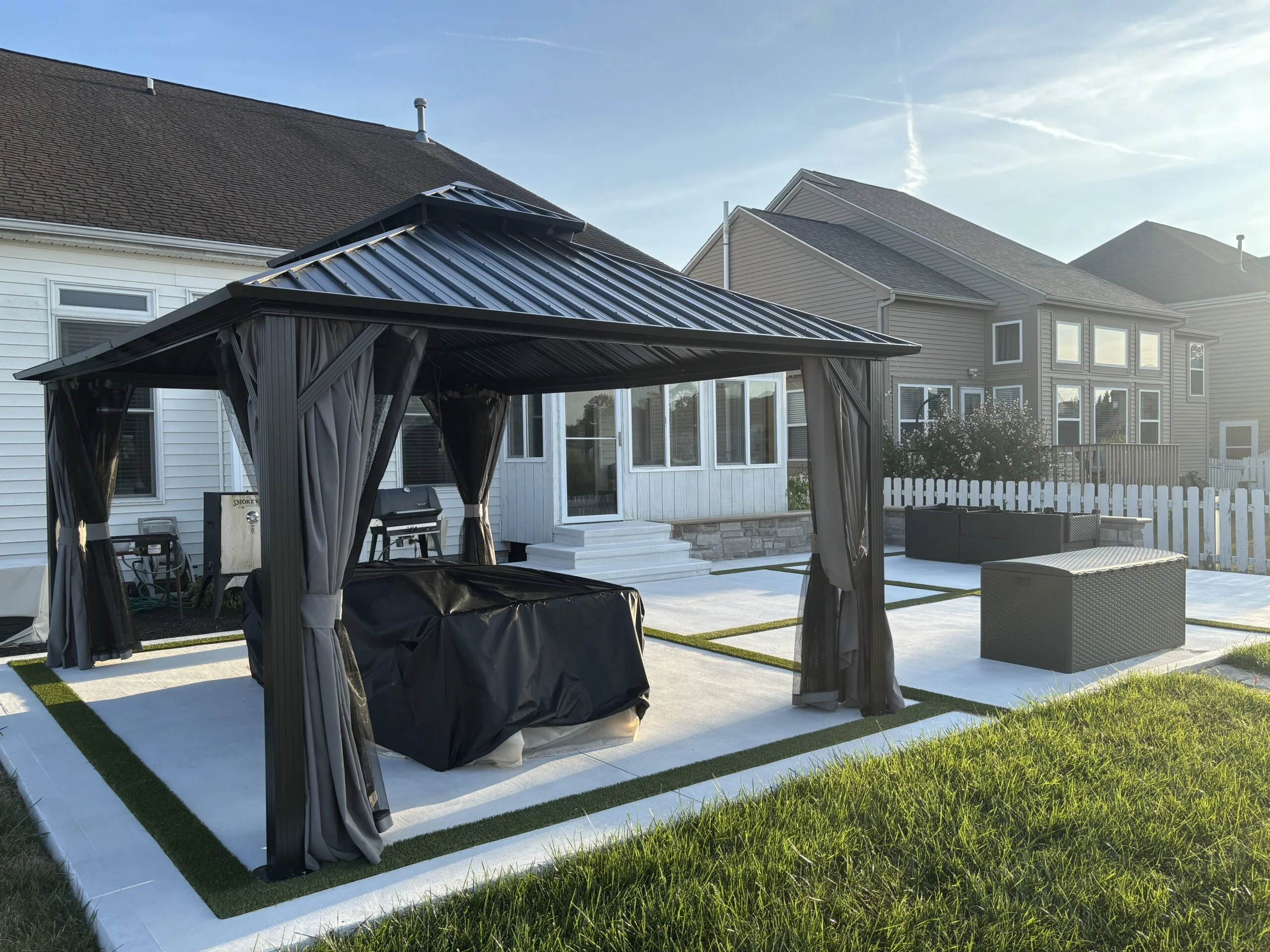Outdoor seating area under a black metal gazebo with gray curtains, white house with large windows in the background on a sunny day.