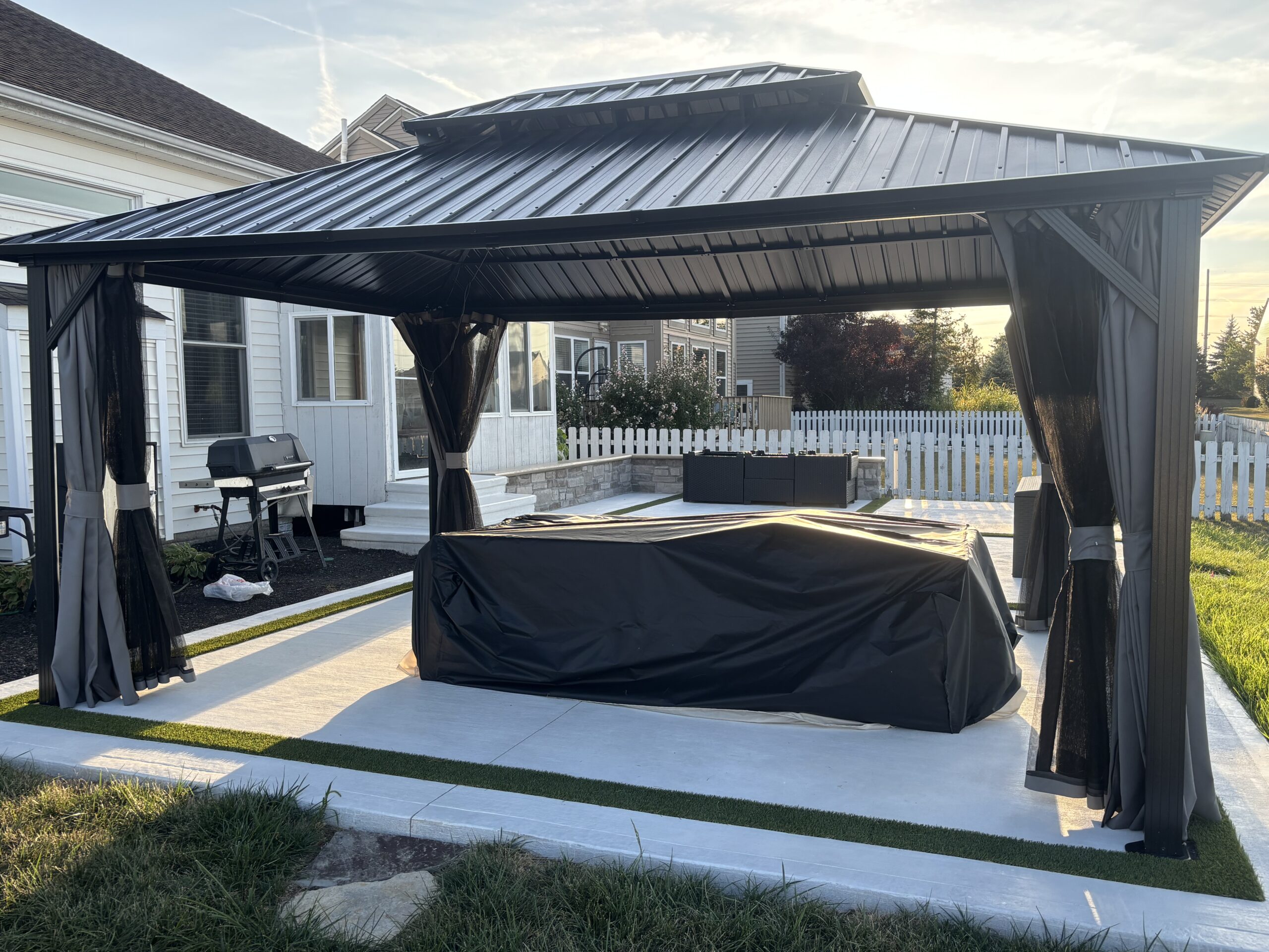 Black metal outdoor pavilion with flowing curtains on a concrete patio, in a backyard beside a white house and grill area