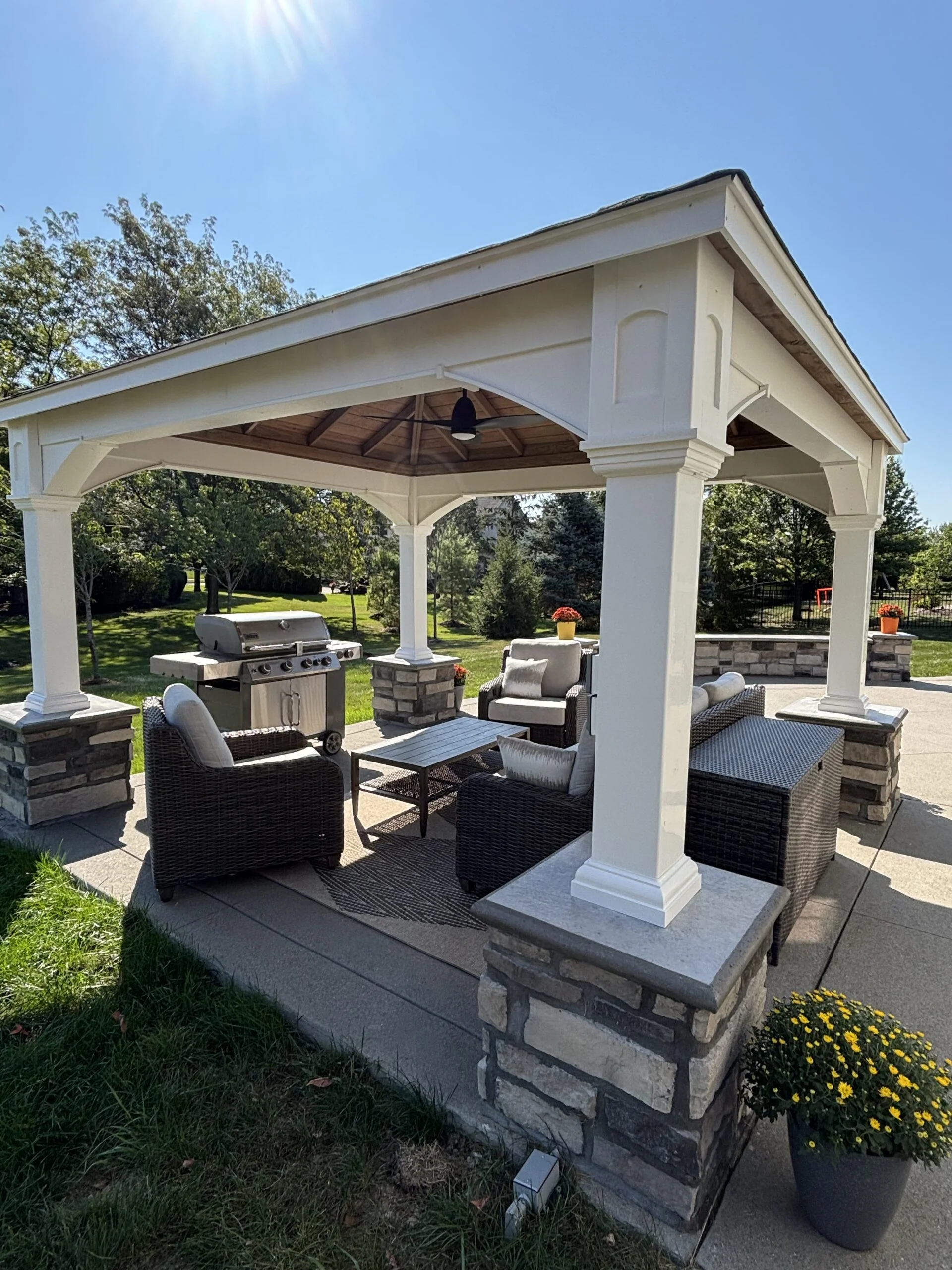 A white outdoor pavilion with stone pedestals, wicker seating, and a built-in grill in a sunny backyard garden.